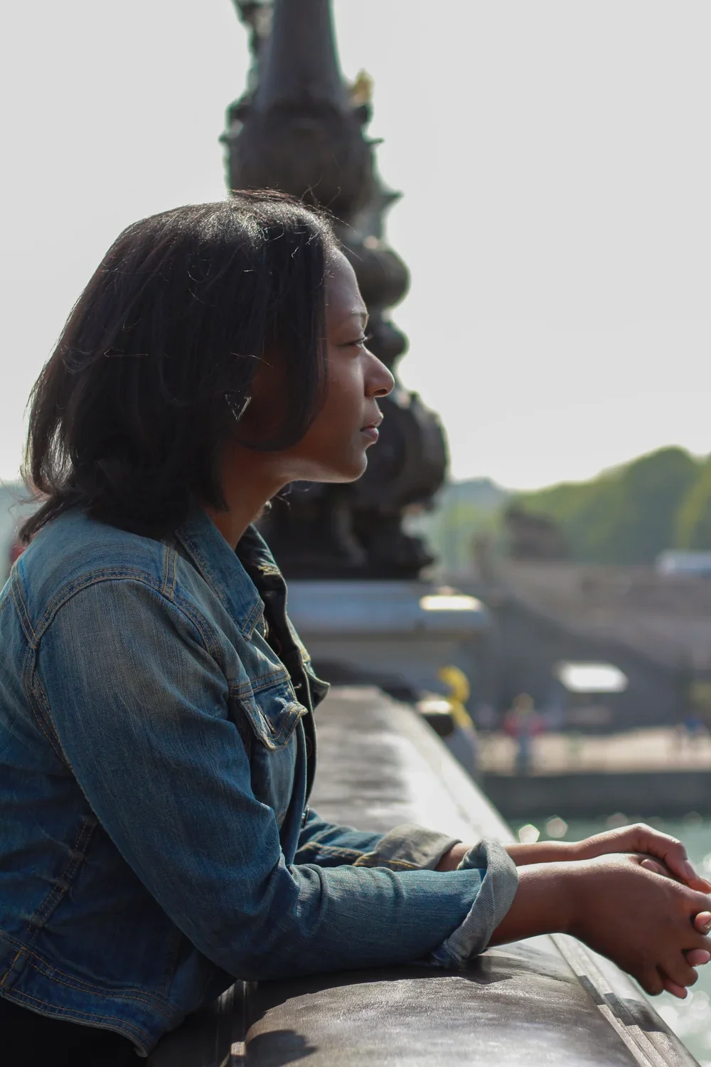 Robin Allison Davis: A woman with dark hair in a denim jacket looking out over a bridge or river with a statue in the background during daylight.
