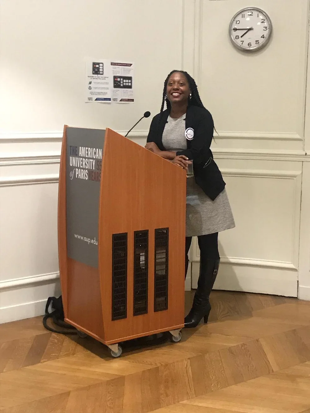 A woman standing at a podium with a microphone, smiling, in a conference room. There is a clock showing 8:44 on the wall behind her and posters on the wall.