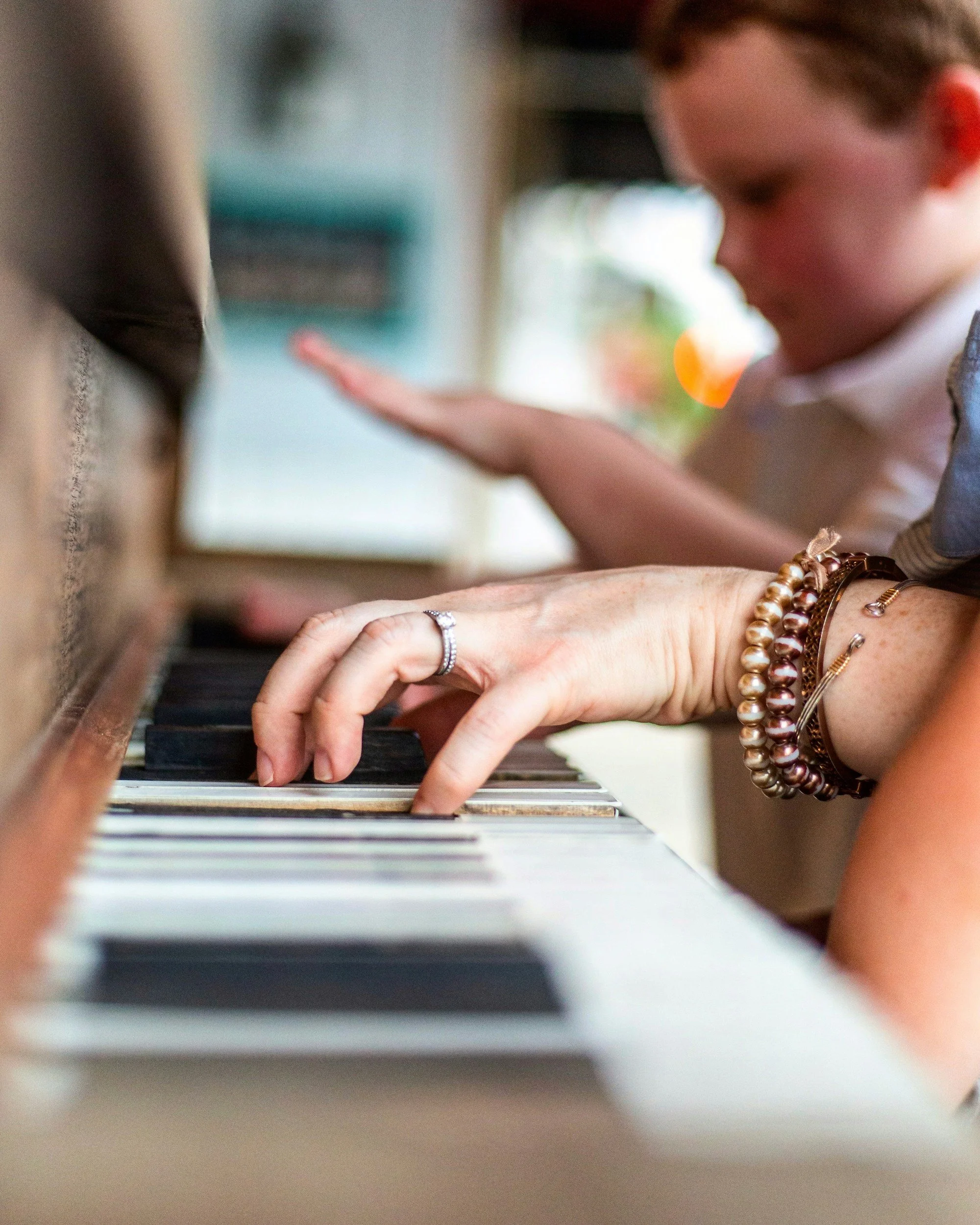 Close-up of a woman's hand with bracelets and a ring playing the piano, with a young boy blurred in the background.