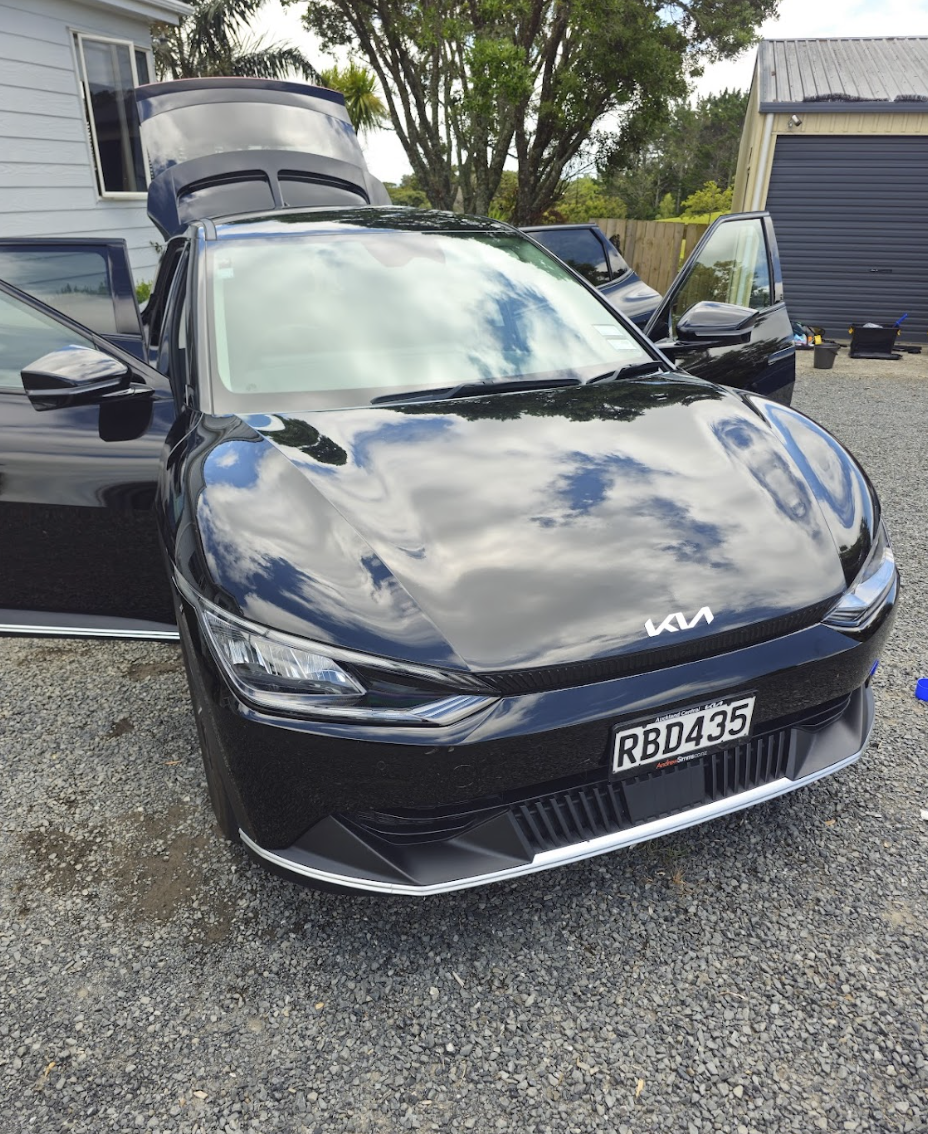 Black Kia car with open doors and hood parked on gravel driveway next to a white house and a garage, reflecting the cloudy sky and trees.