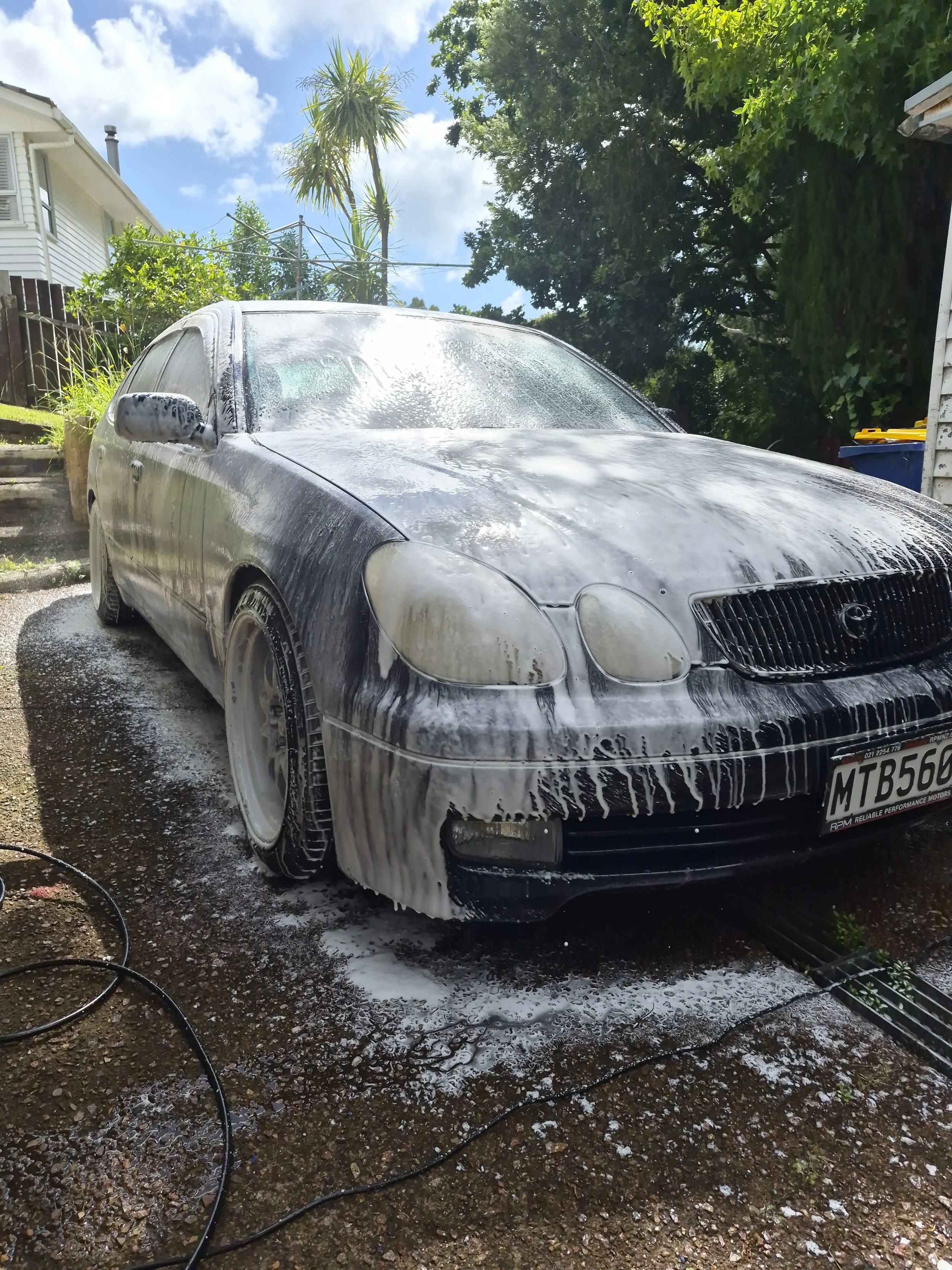 A black car covered in white soap suds being washed with water, with soap foam dripping off the car and soap suds on the ground, in an outdoor setting with trees and a blue sky with clouds.