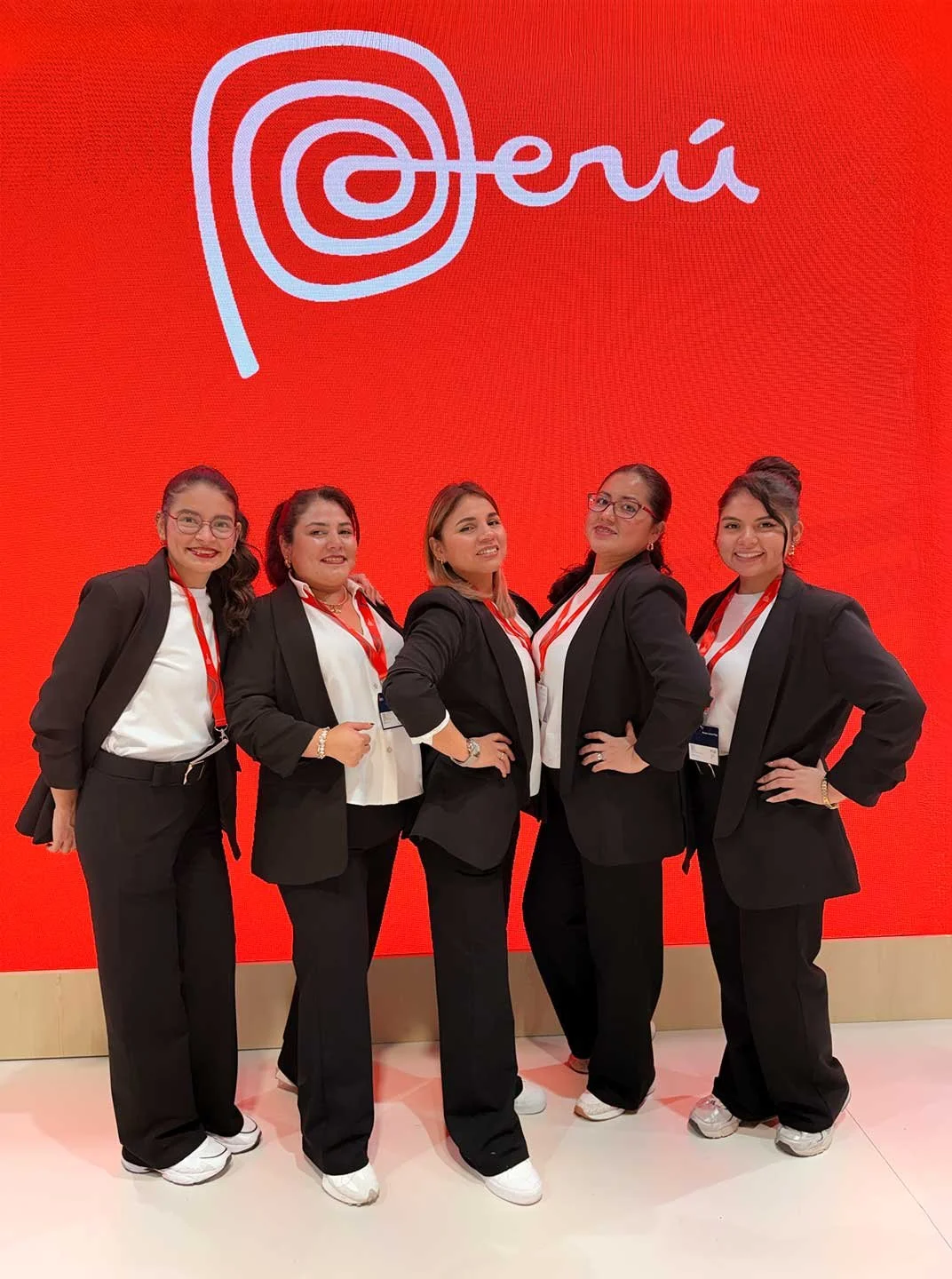 Cinco mujeres en trajes formales con gafetes, posando frente a un fondo rojo con la palabra 'Perú' escrita en letra cursiva y un diseño en espiral