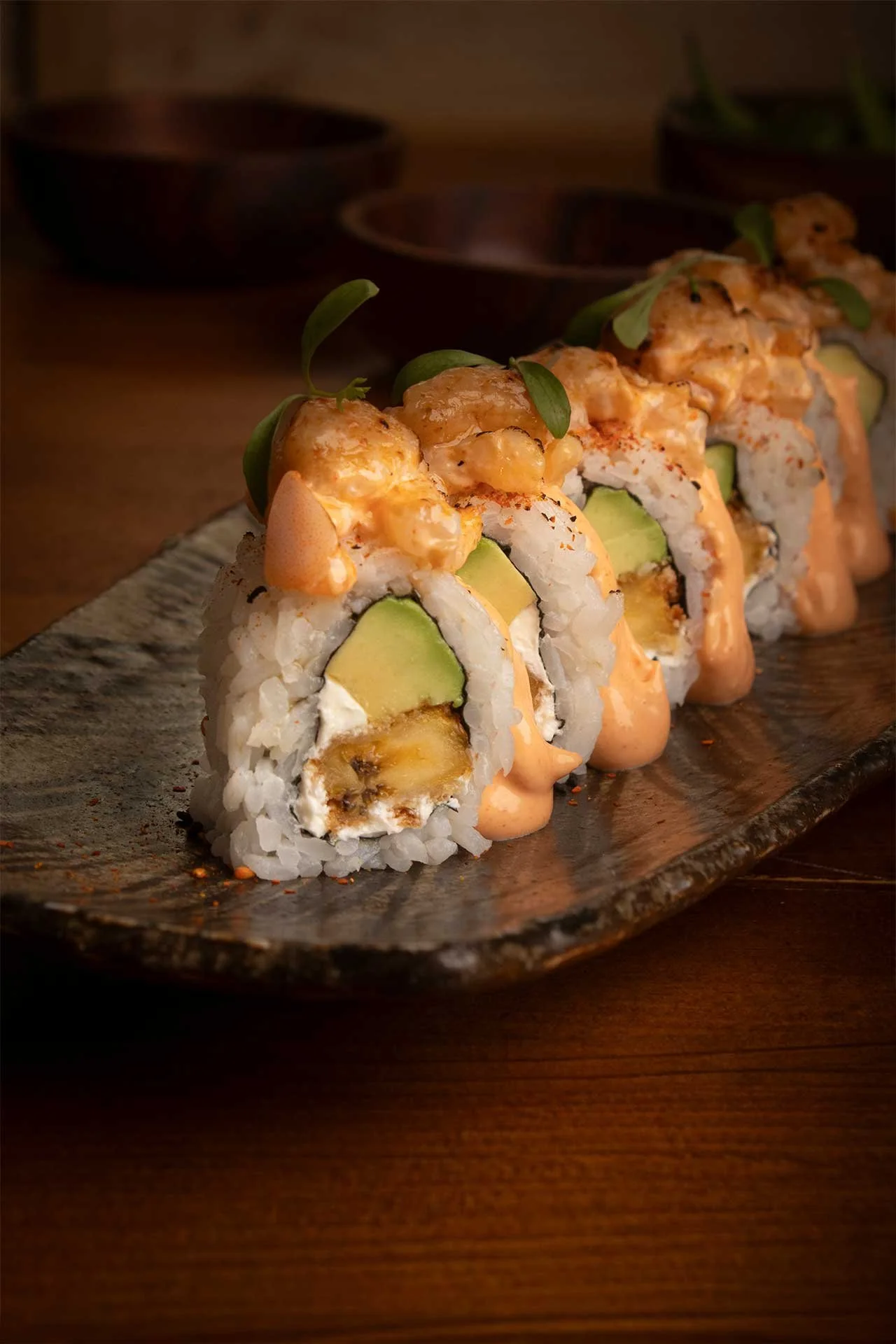 Rollos de sushi con aguacate, cangrejo y salsa, decorados con hojas verdes y servidos en un plato de cerámica sobre una mesa de madera.