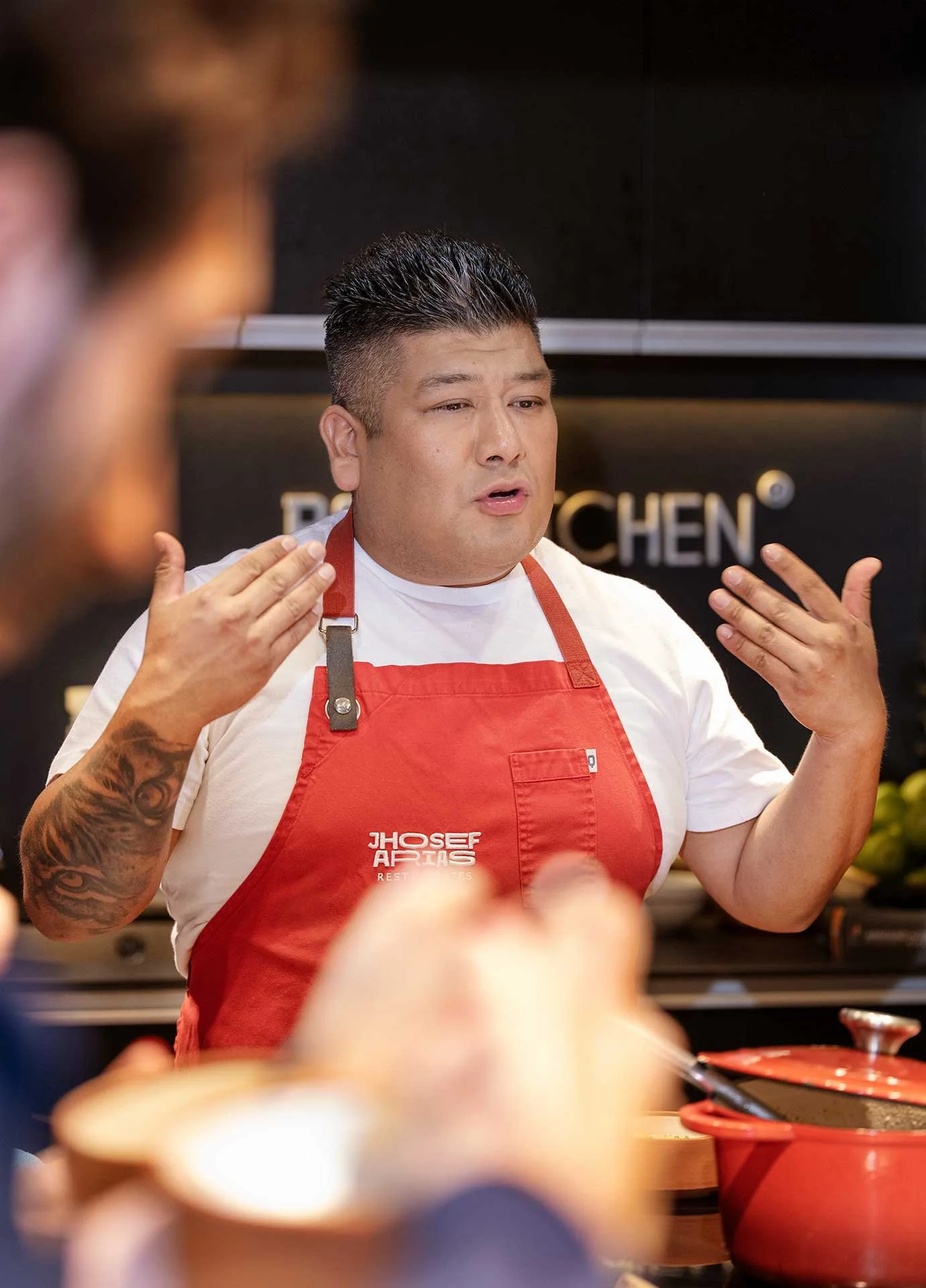 Chef profesional explicando en cocina, lleva delantal rojo y camiseta blanca, con tatuajes en el brazo, en un entorno de cocina moderna.
