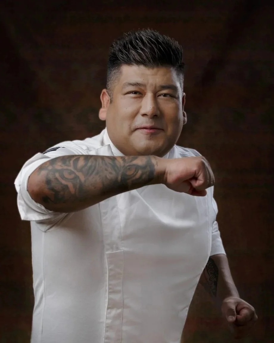 Hombre con peinado moderno y tatuajes en el brazo, vestido con uniforme de chef, haciendo un gesto de fuerza con el puño. Fondo oscuro.