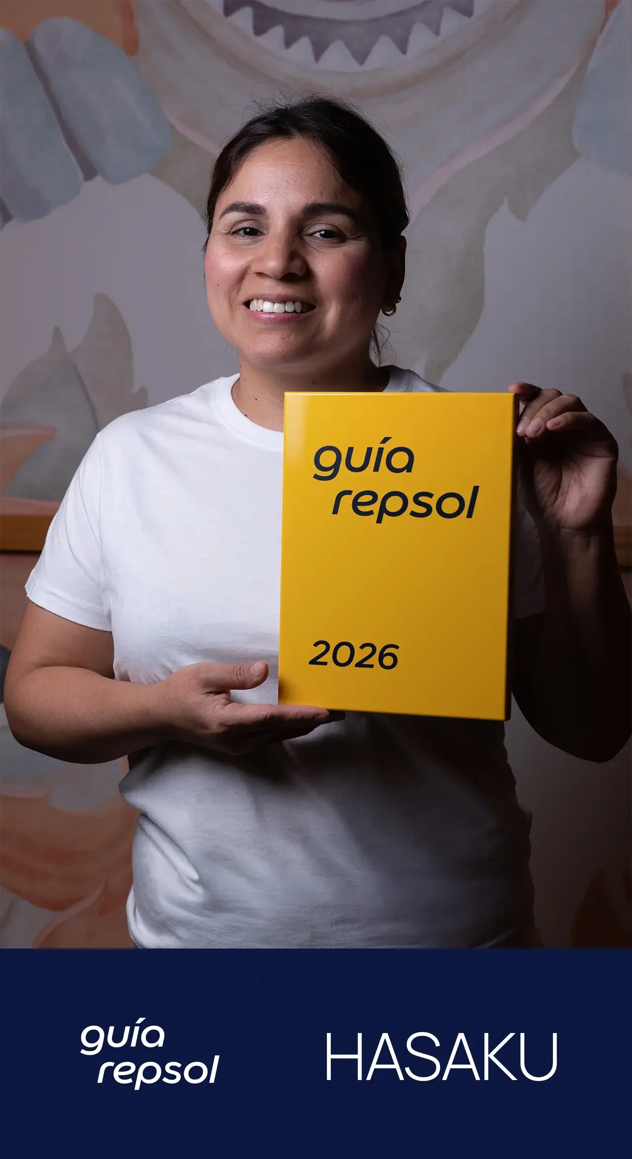 Una mujer de cabello castaño oscuro y rostro sonriendo sostiene una guía amarilla con texto en negro que dice 'guía repsol 2026'.