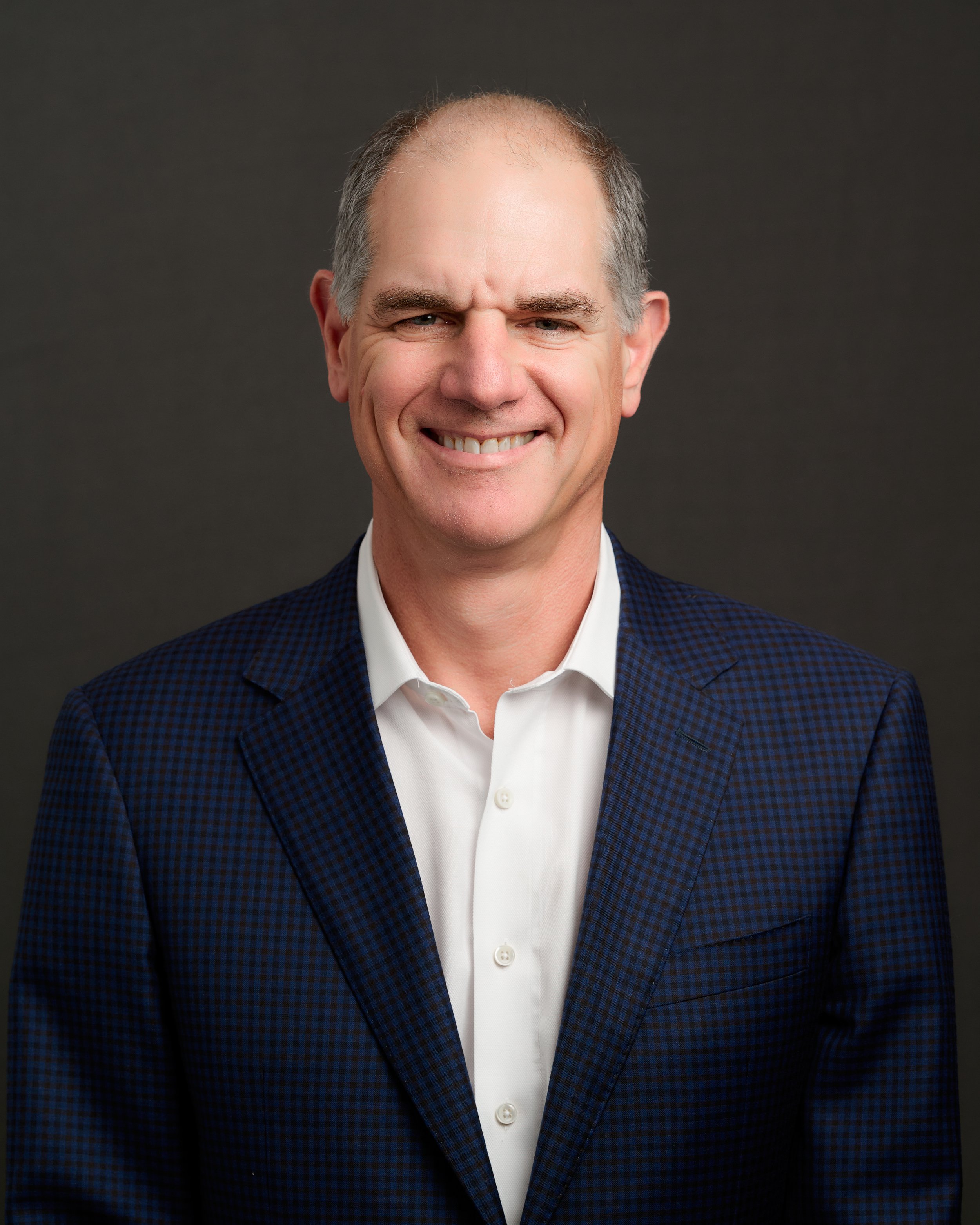 A middle-aged man with gray hair, smiling, wearing a white shirt and a dark blue checkered blazer, against a dark background.