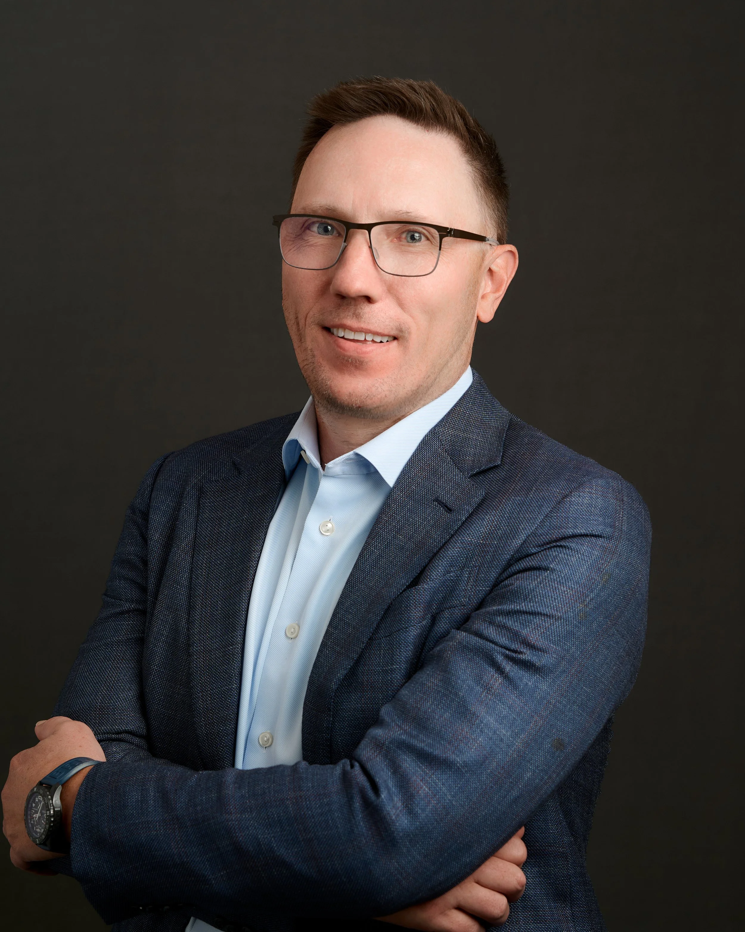 A professional man in a suit and glasses smiling against a dark background.