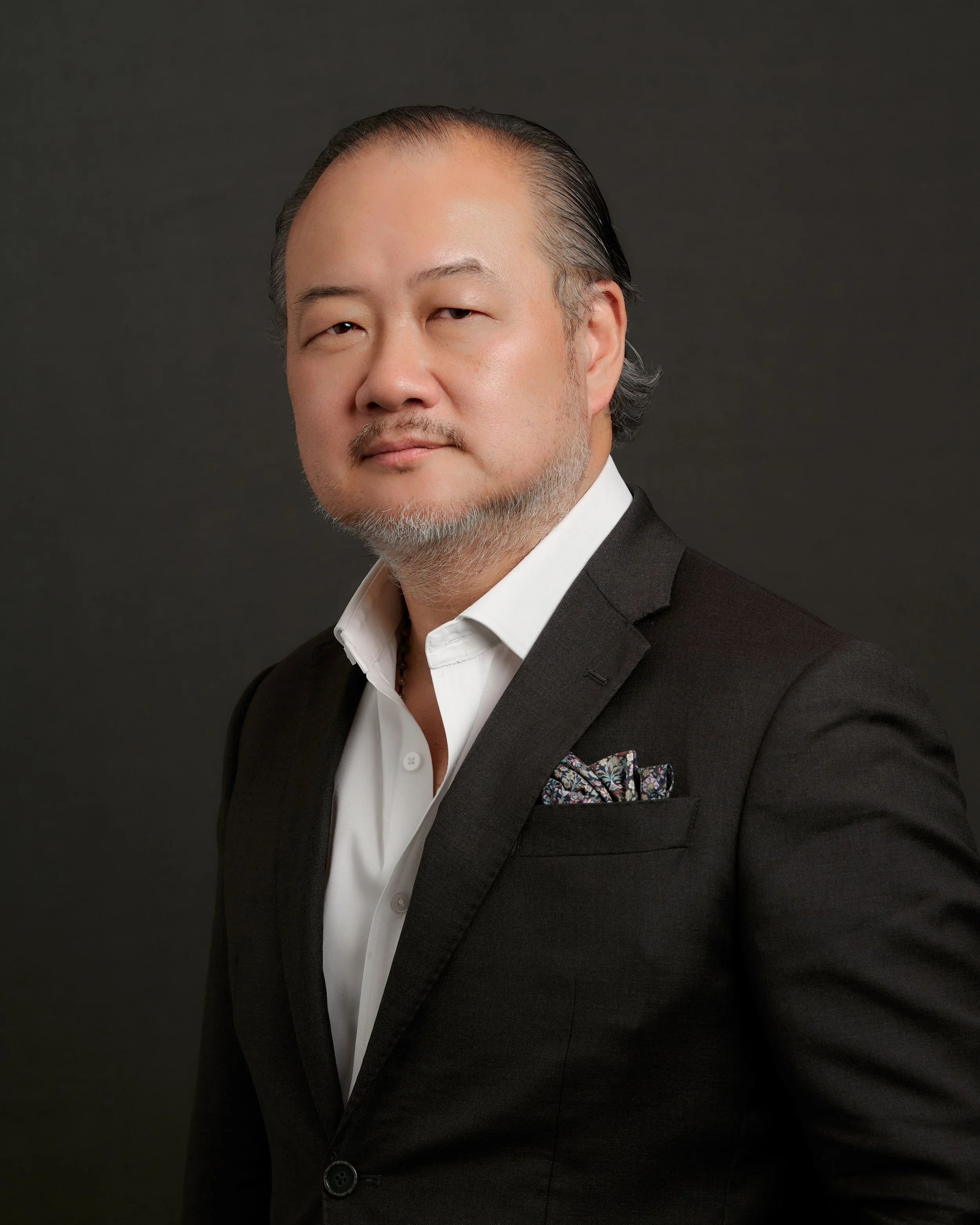 Portrait of a middle-aged man with a beard, wearing a black suit, white shirt, and a patterned pocket square, against a dark background.