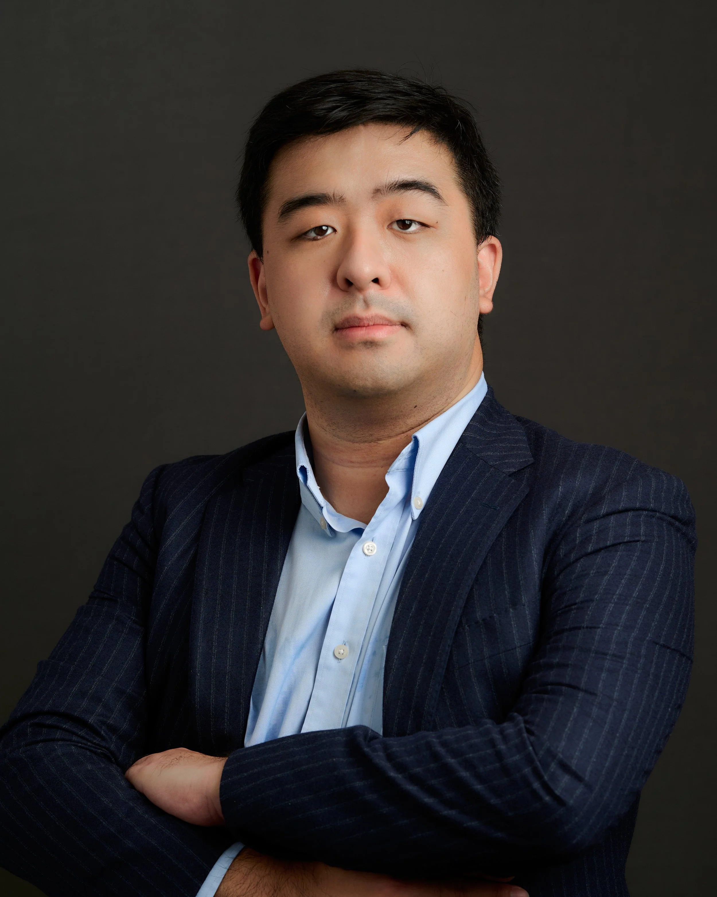 A professional headshot of a man in a dark suit with crossed arms against a dark background.