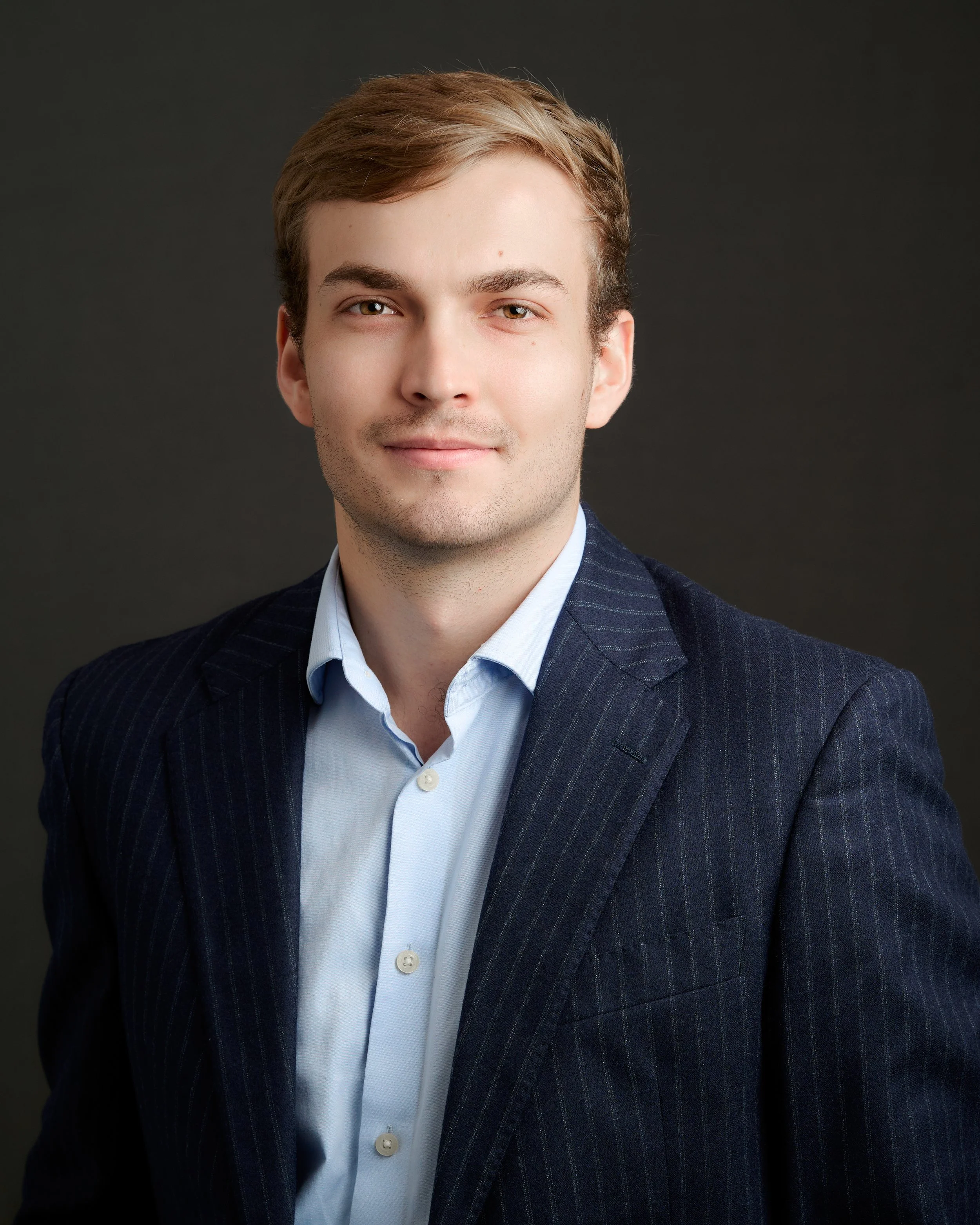 Portrait of a young man with light brown hair, wearing a dark pinstripe suit jacket and a light blue shirt, against a dark background.