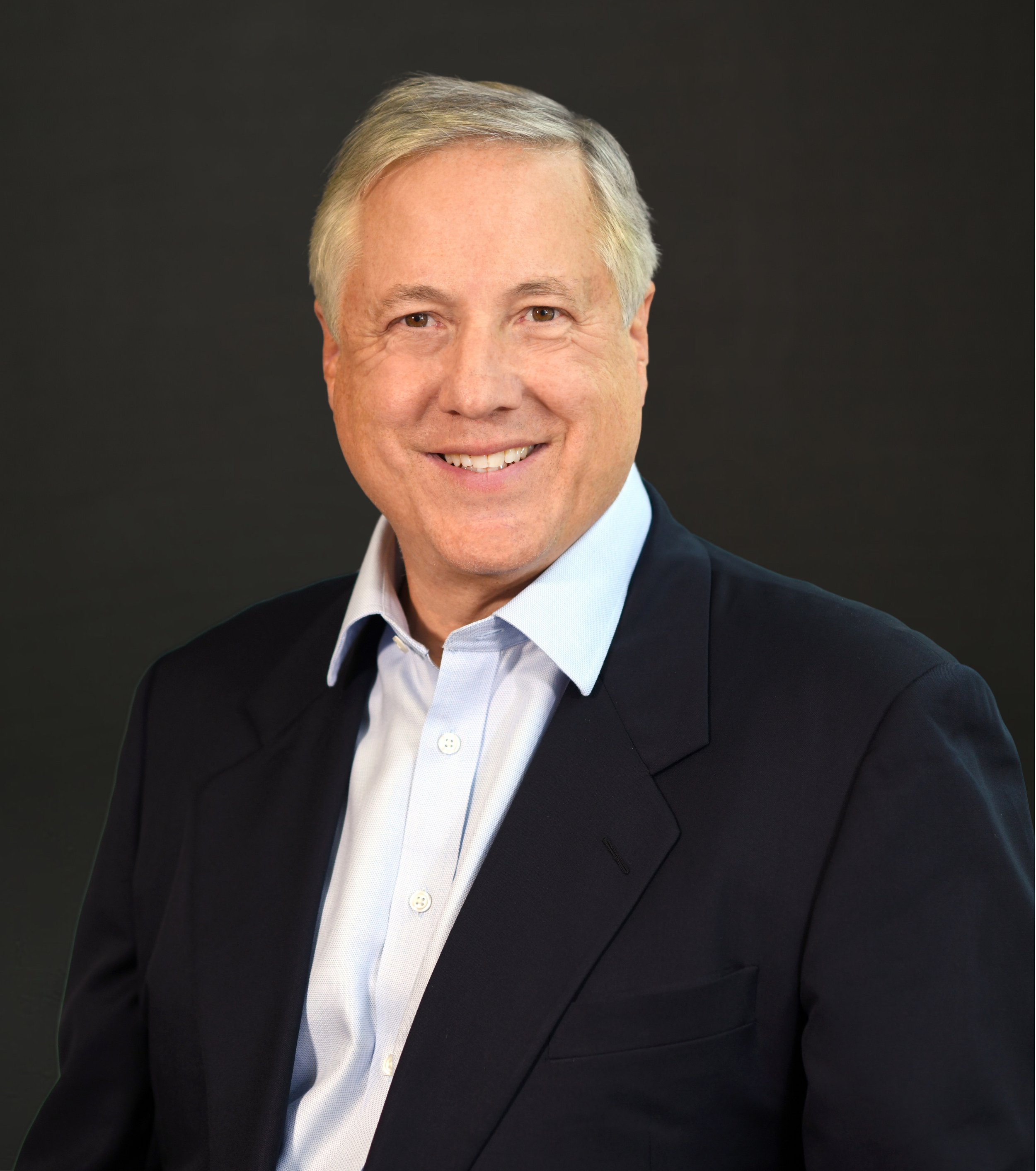 A middle-aged man with gray hair wearing a navy blazer and light blue collared shirt, smiling against a black background.