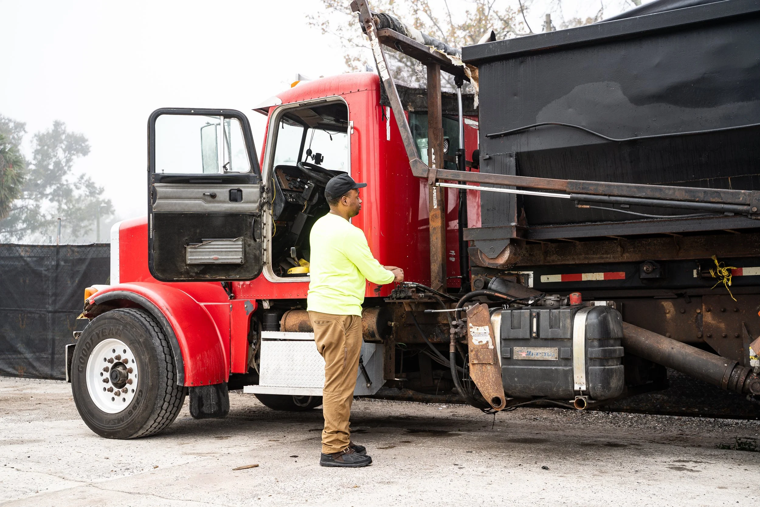 A man in a yellow shirt and khaki pants standing beside a large red dump truck, inspecting or working on the truck's engine or controls.