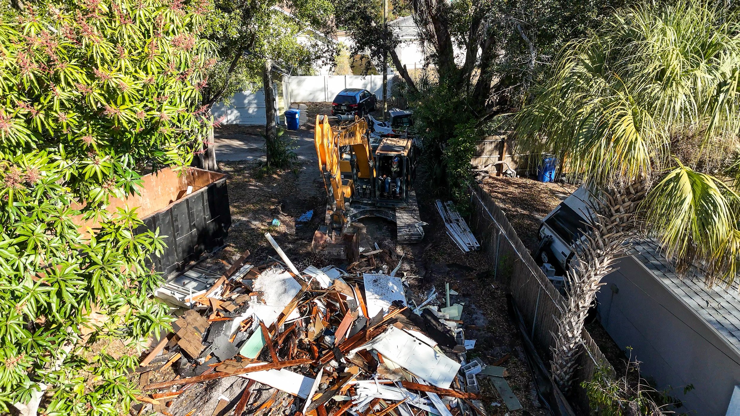A construction site with a yellow excavator tearing down a building, debris scattered on the ground, and a palm tree and a large shrub nearby, with parked vehicles and trees in the background.