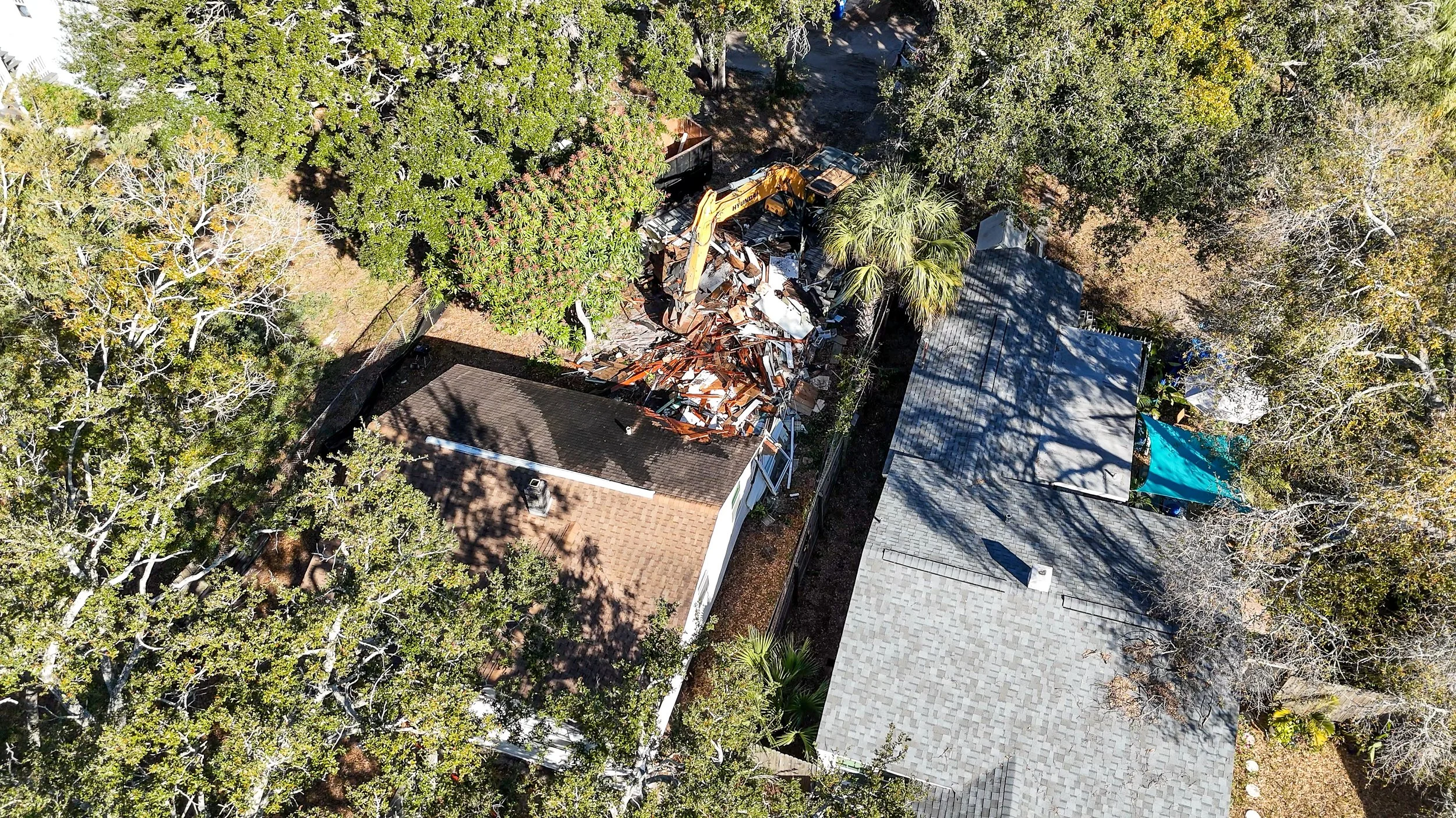 Aerial view of a house with damaged roof debris, a construction excavator, and trees surrounding the property.