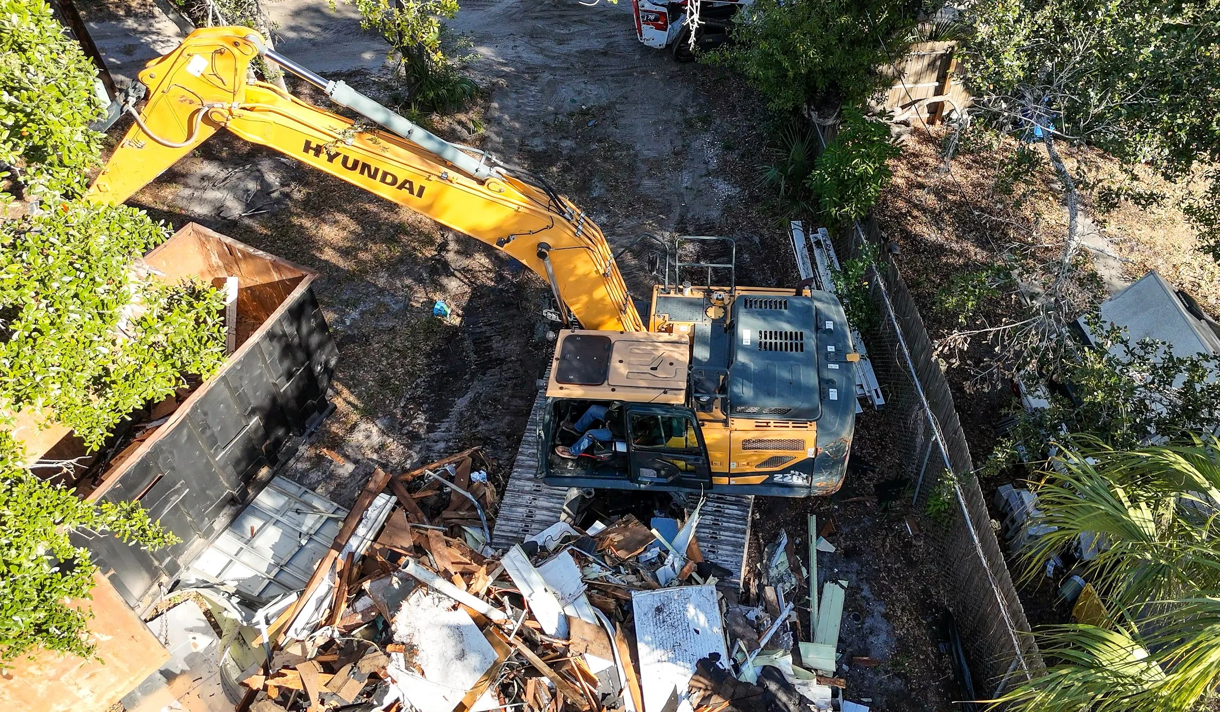 A yellow Hyundai excavator demolishing a wooden structure with debris, surrounded by trees and a fence.