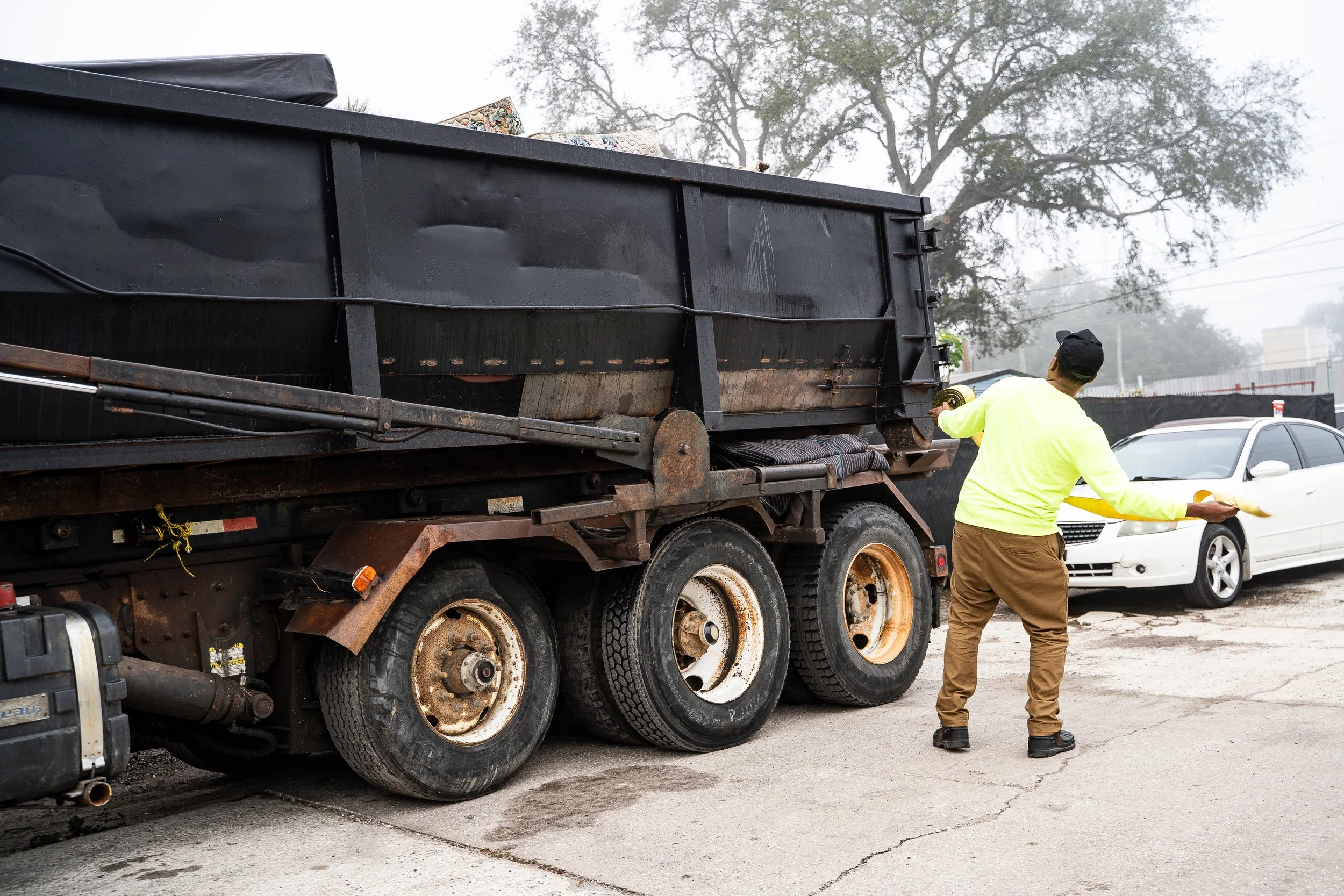 A person wearing a high-visibility yellow jacket and brown pants holding a tape measure, standing next to a large black truck and a white car in an outdoor lot with cloudy sky and trees in the background.