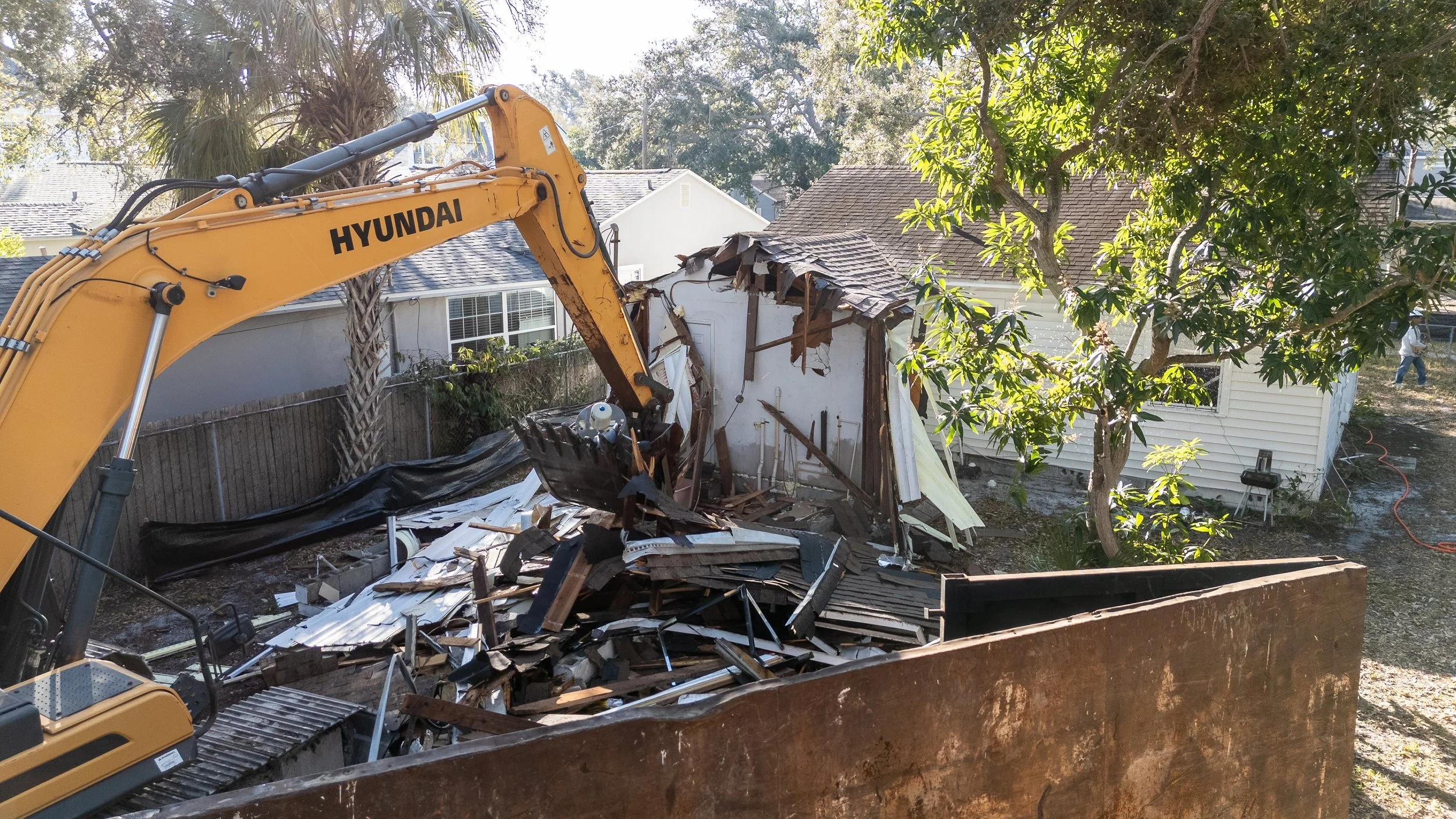 A yellow Hyundai excavator demolishing a small white house with partial roof and damaged walls, in a residential neighborhood with trees and neighboring houses visible.