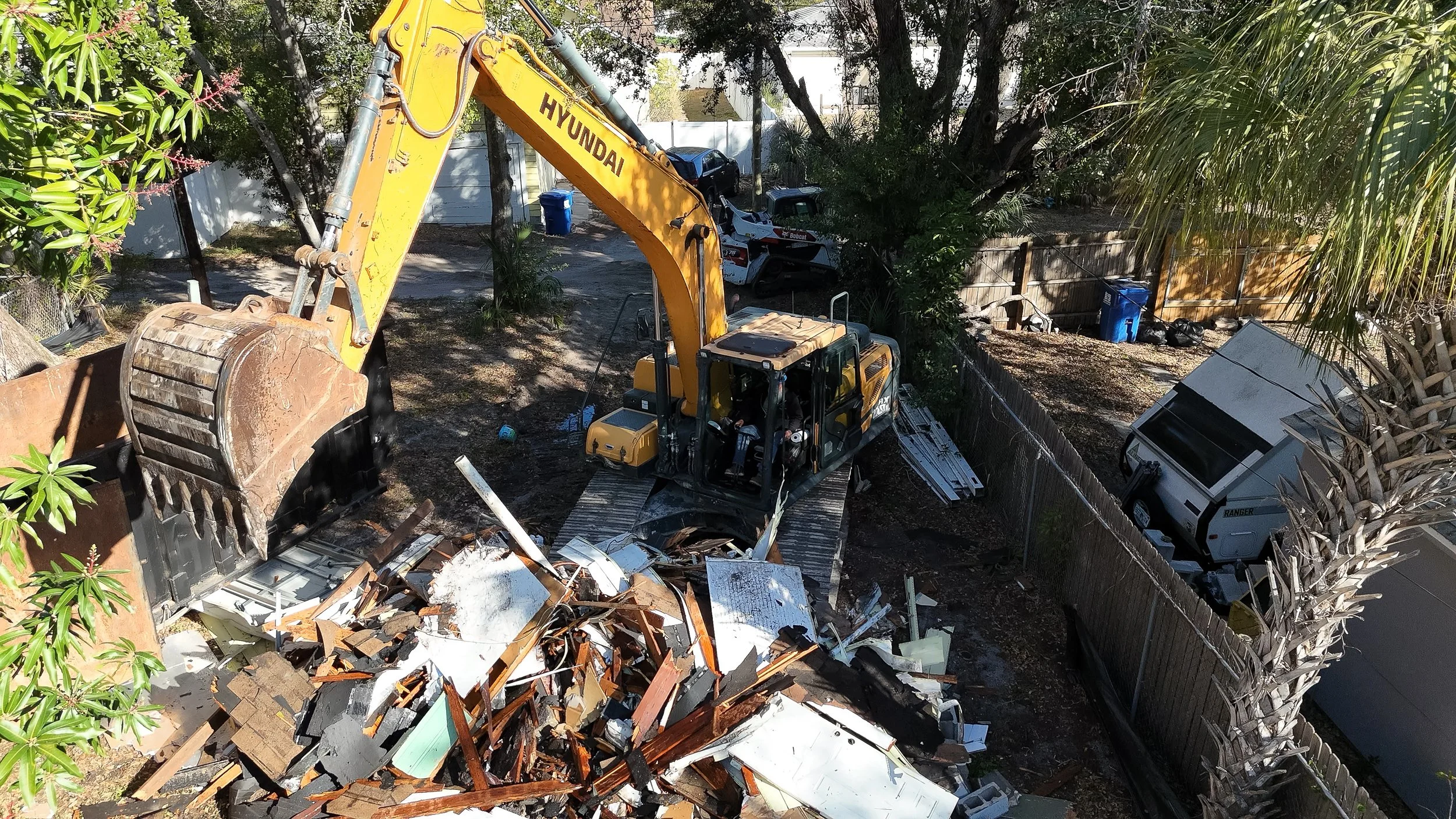 Yard with construction excavator demolishing a pile of debris, including wood and metal, surrounded by trees, a chain-link fence, and a trash truck in the background.
