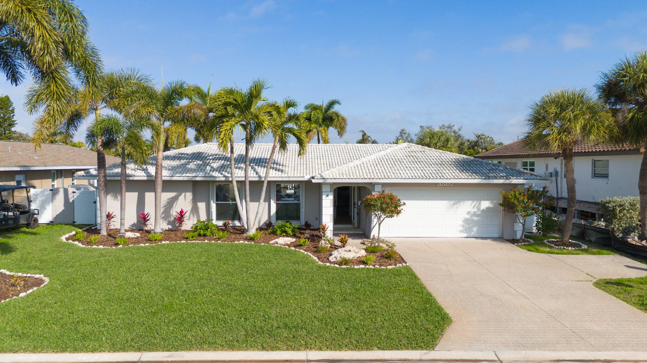 A modern single-story house with a white exterior, white tiled roof, a two-car garage, and a manicured lawn with tropical plants and palm trees in a suburban neighborhood.