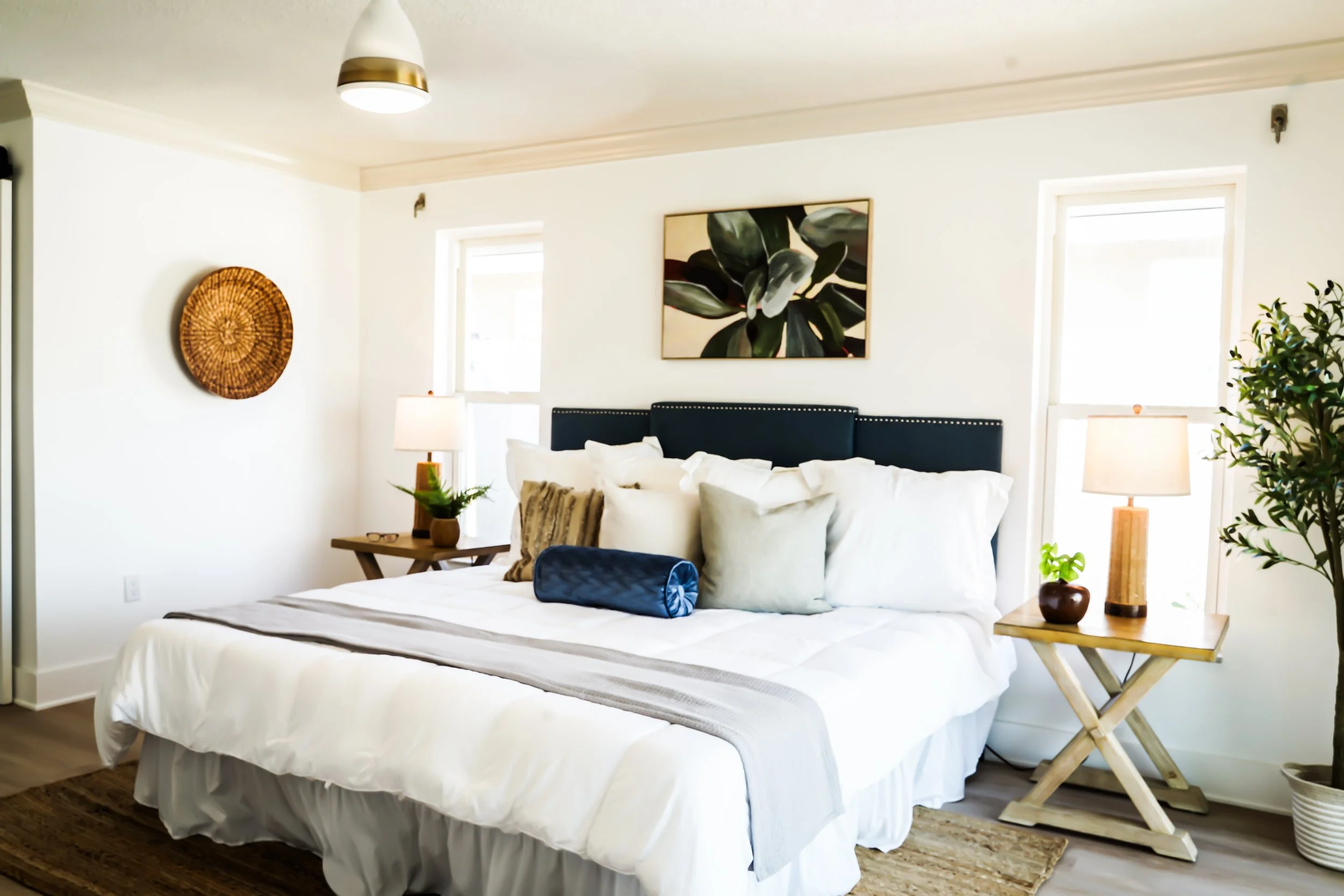 Bright bedroom with a large bed, white bedding, multiple pillows, navy headboard, two wooden side tables with lamps, artwork of green leaves on the wall, potted plants, and natural light from windows.