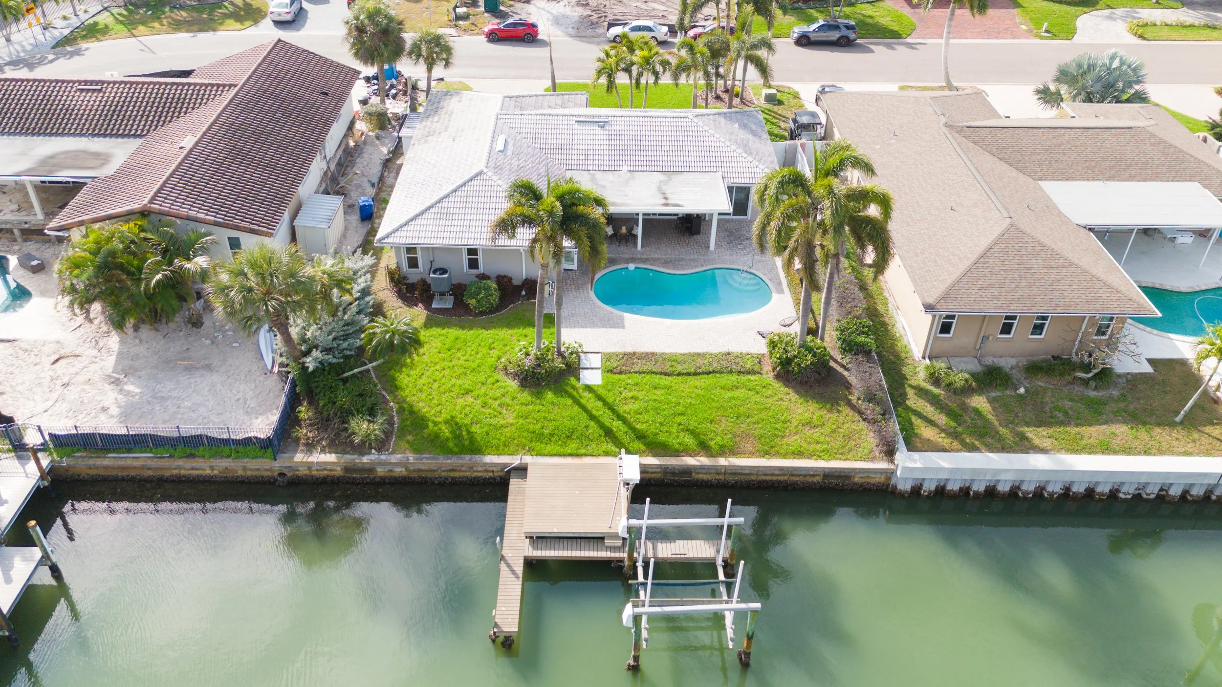Aerial view of a suburban neighborhood with three houses along a canal. The middle house has a backyard with a small swimming pool, palm trees, a lawn, and a dock with boat lift on the water.