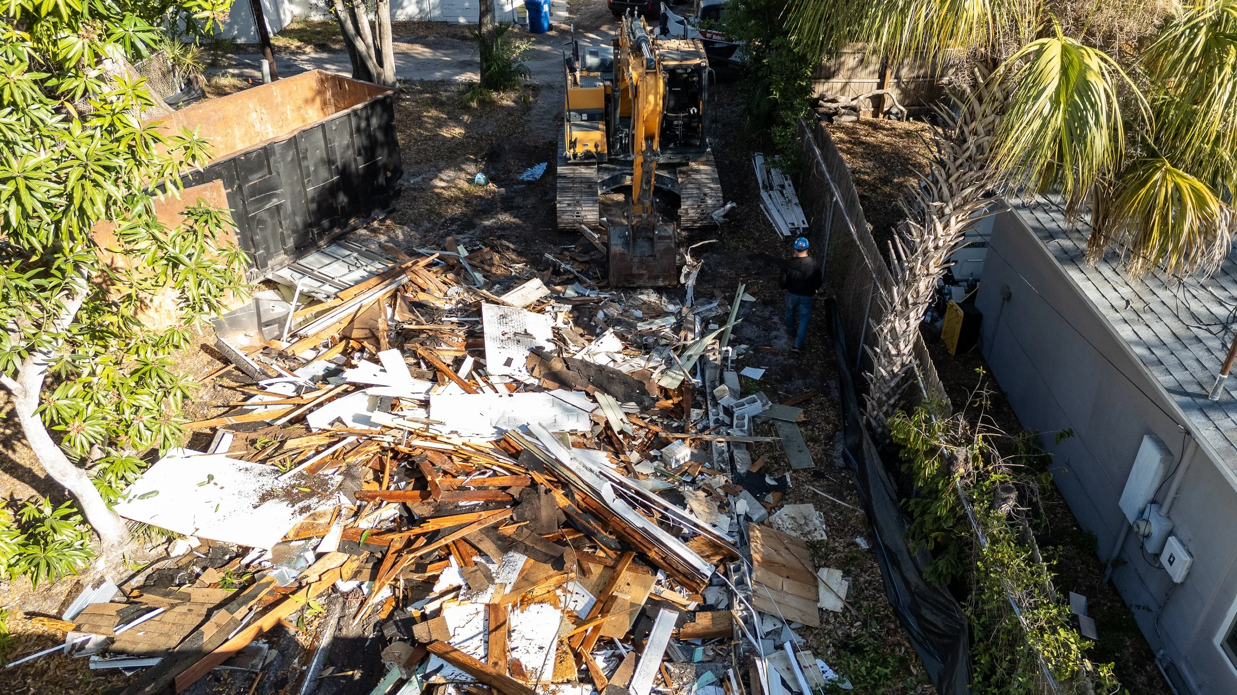An aerial view of a backyard construction or demolition site with debris, a small excavator, and two workers, one wearing a blue helmet and the other wearing a black cap, standing near a fence with trees and a building on the sides.