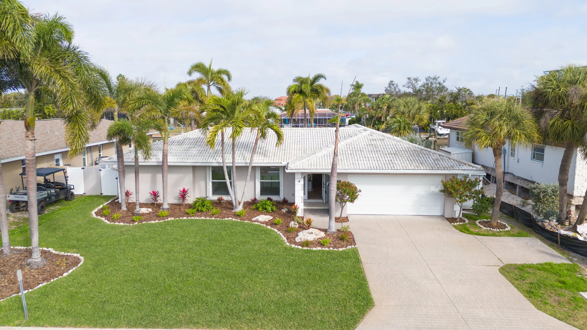 Front yard view of a single-story house with white exterior, tiled roof, and a two-car garage, surrounded by landscaped lawn with palm trees and decorative plants