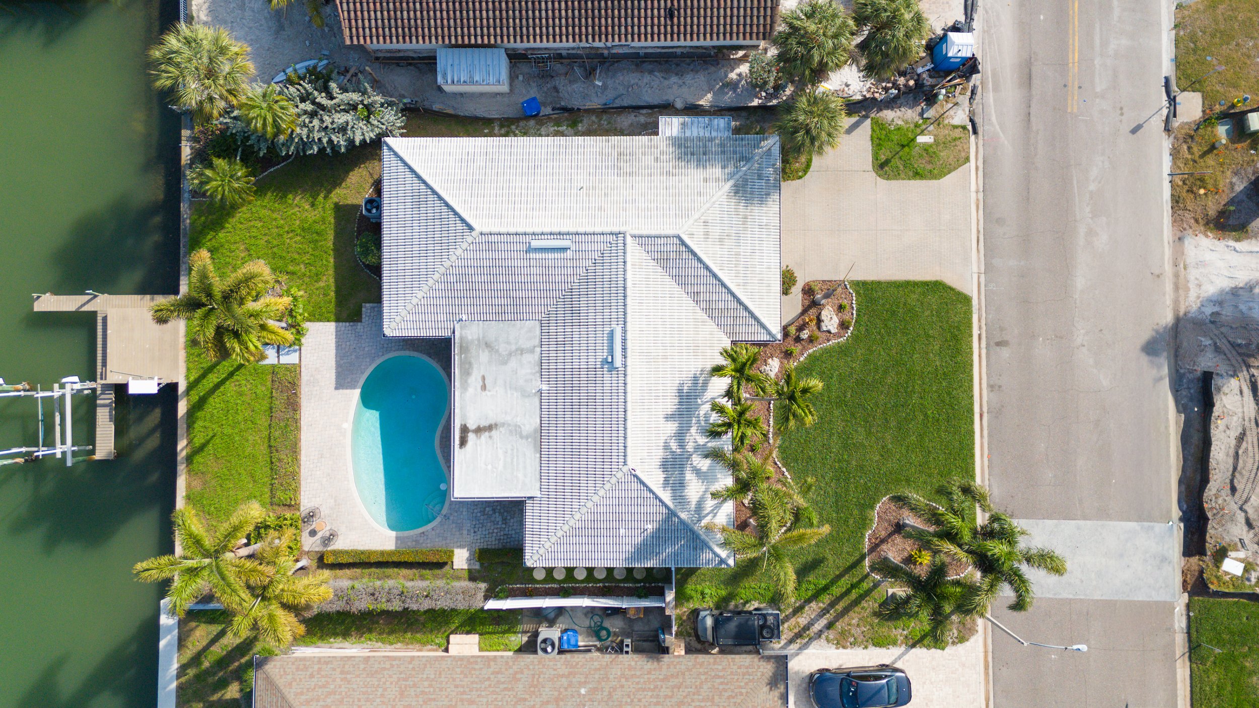 Aerial view of a house with a swimming pool, palm trees, a green lawn, and a driveway, next to a canal with a dock on the left side.