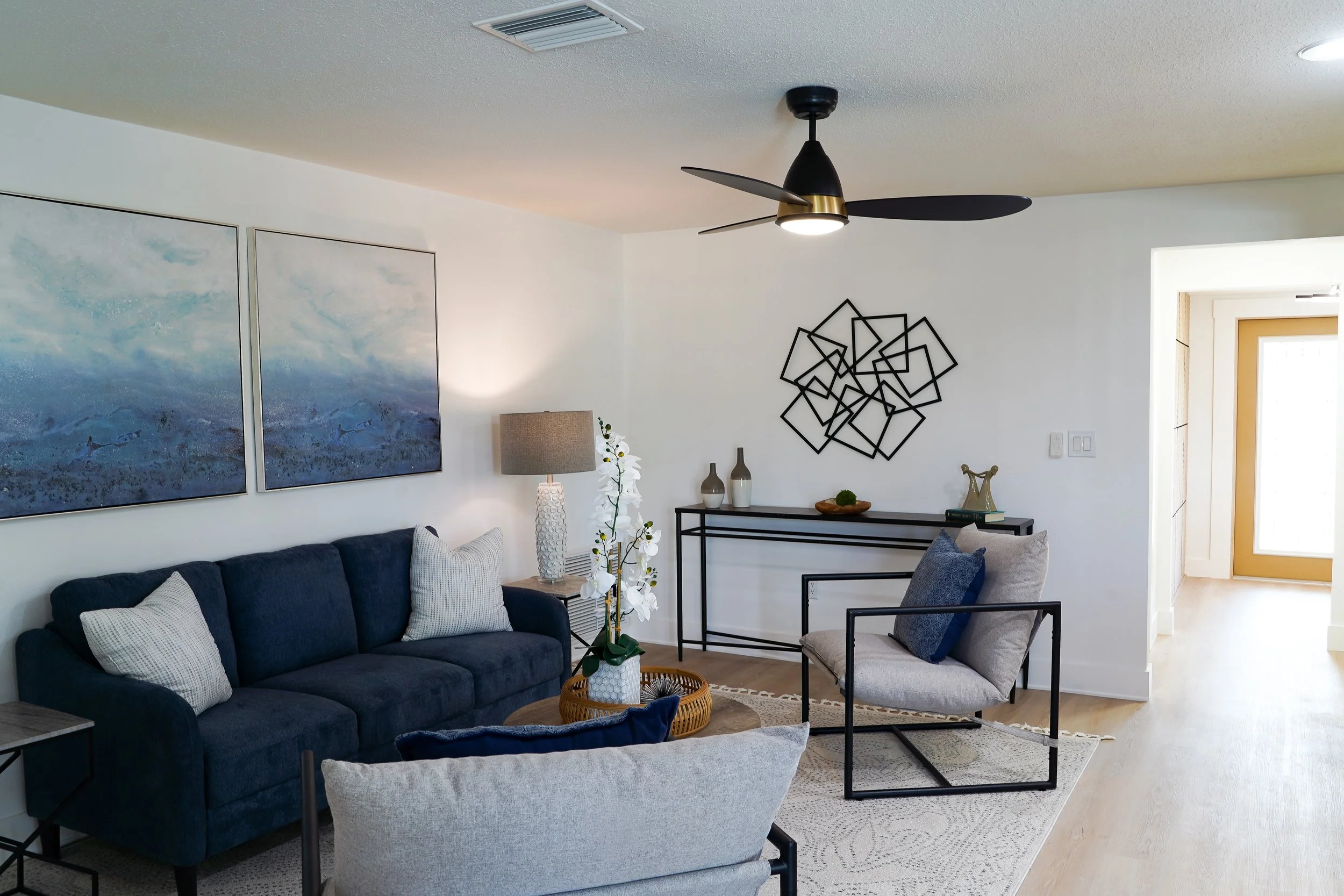 Living room with a navy blue couch, a beige armchair, a modern black metal and glass console, a decorative black wall art, a floor lamp, a floral arrangement, and wall art in a bright, minimalist space.
