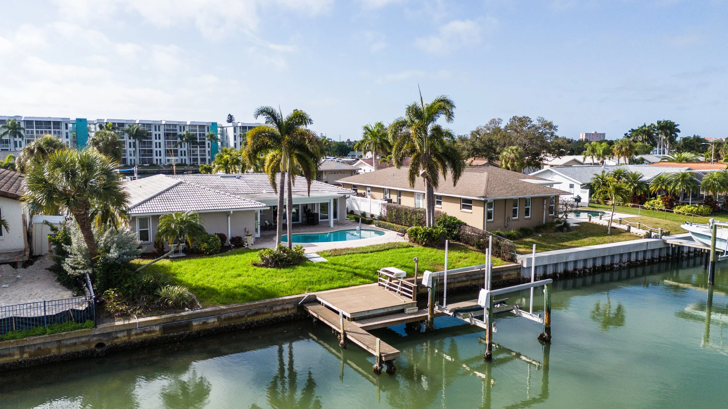 Residential houses along a waterway with a dock, palm trees, and a swimming pool under a partly cloudy sky.