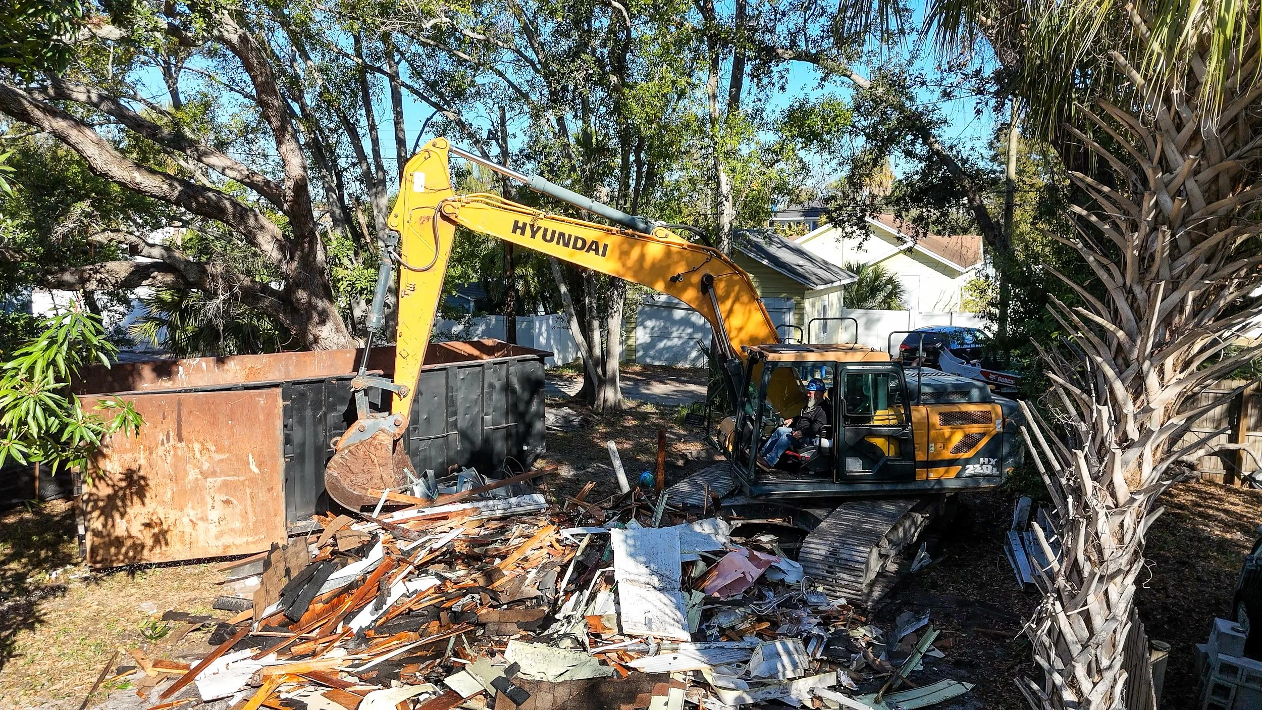 A yellow Hyundai excavator clearing debris in a backyard surrounded by trees and a fence.