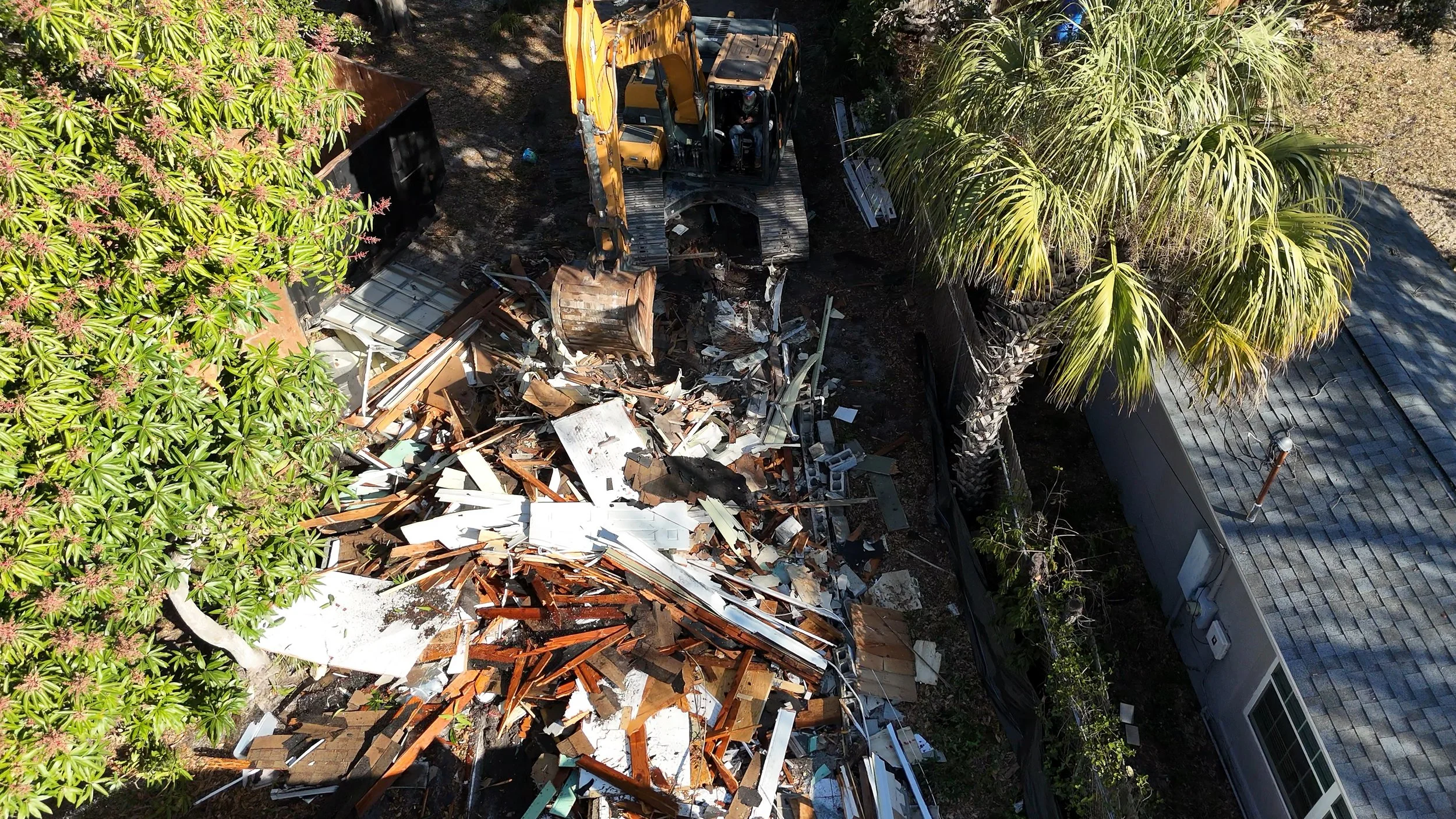 A construction excavator removing debris from a demolished building in a backyard with trees and a house with a gray roof.