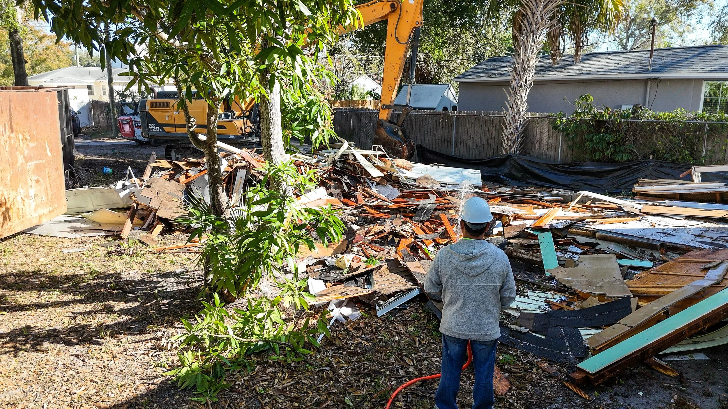 A man wearing a gray hoodie, jeans, and a white helmet stands in front of a pile of debris from a demolished building. A construction excavator is visible in the background along with trees, a house, and a black covering on the ground.
