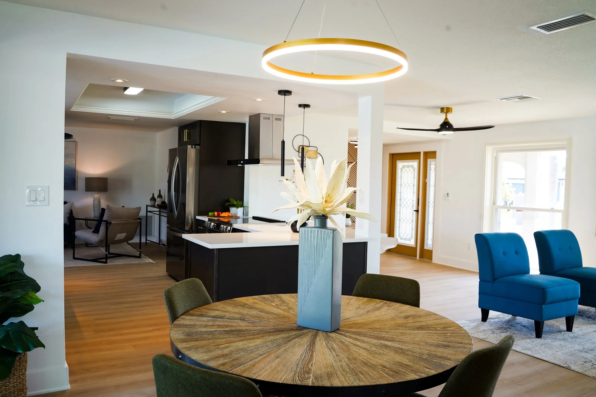 Interior view of a modern home with a round wooden dining table, a large white flower in a gray vase, a kitchen with black cabinets, and a living area with blue chairs, a white wall, and a ceiling fan.