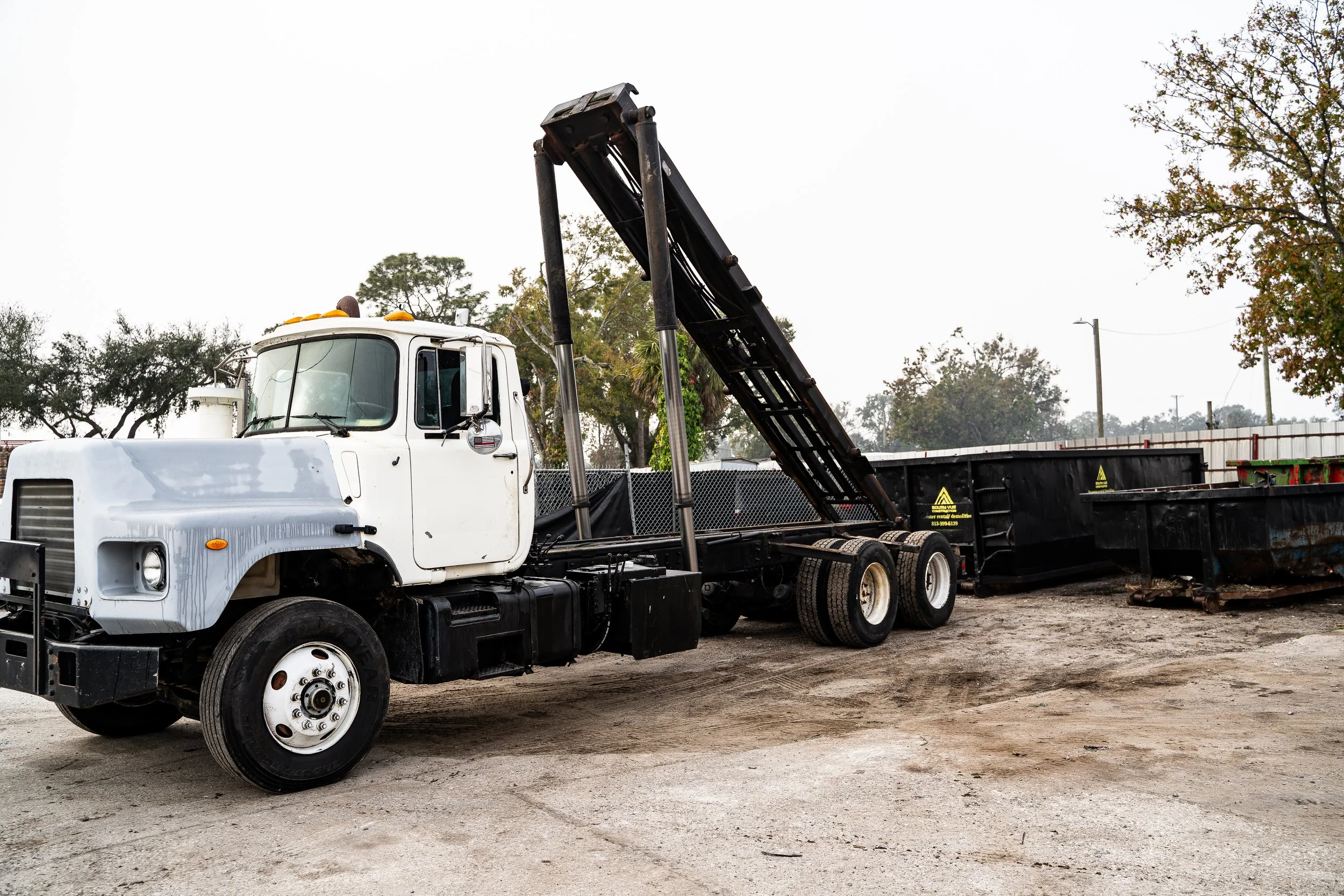 A white tow truck with black flatbed in a dirt lot, with trees and a cloudy sky in the background.
