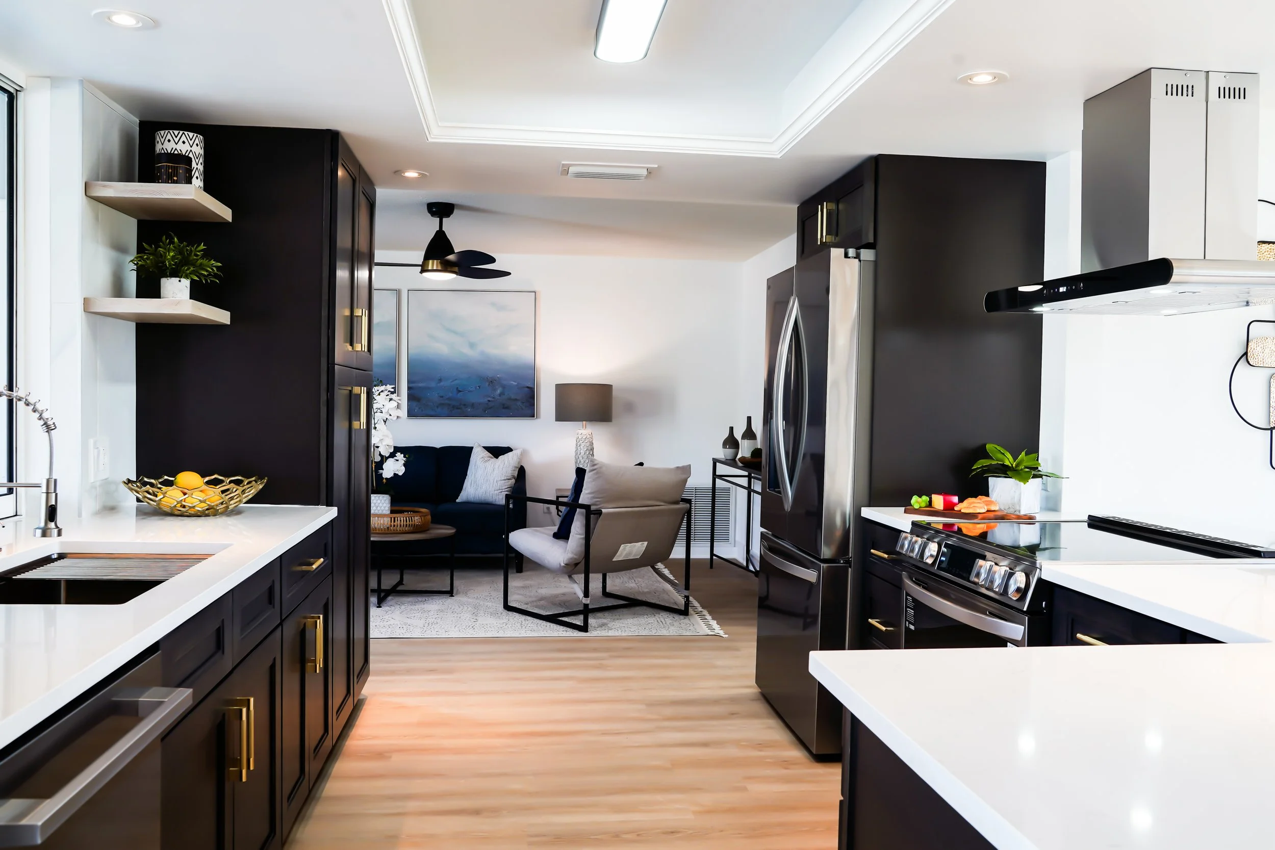 Modern open-concept kitchen with black cabinets and white countertops, adjacent to a living area with a black sofa, white armchair, and abstract blue painting on the wall.