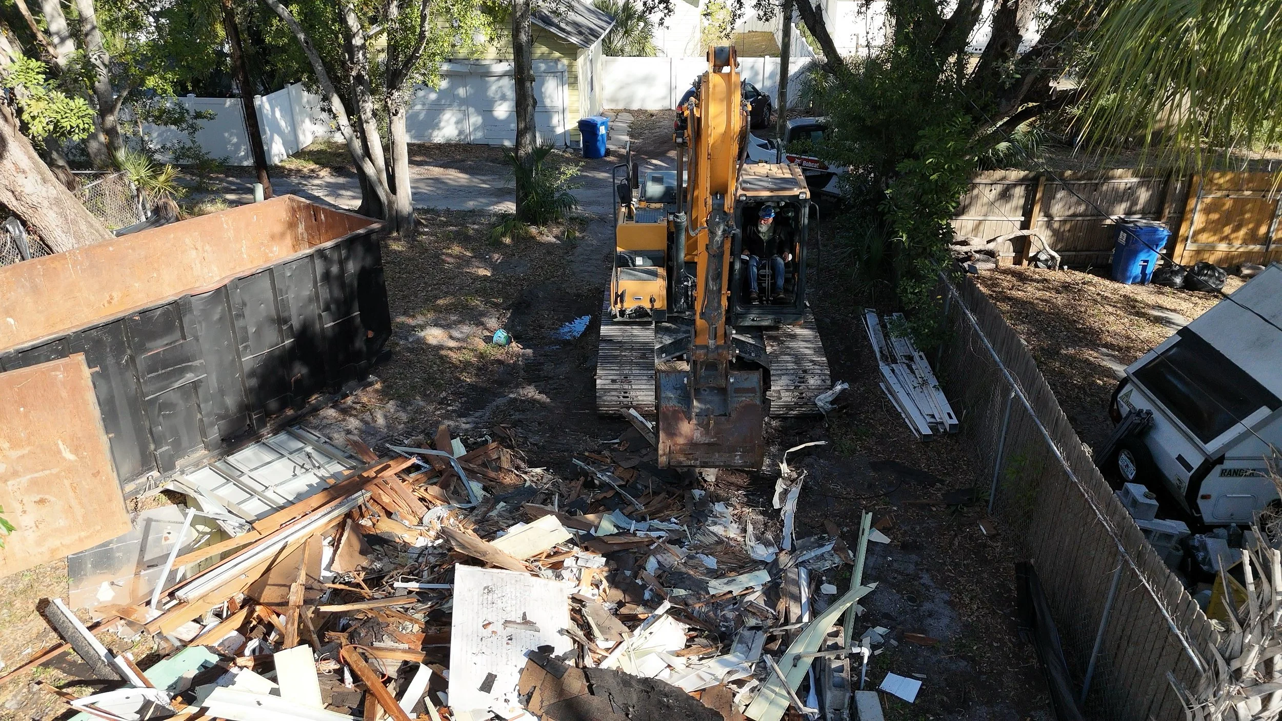 An aerial view of a small construction or demolition site with a yellow excavator moving debris, a toppled large metal container, and scattered construction materials, all enclosed by a wooden and chain-link fence with trees and residential houses in