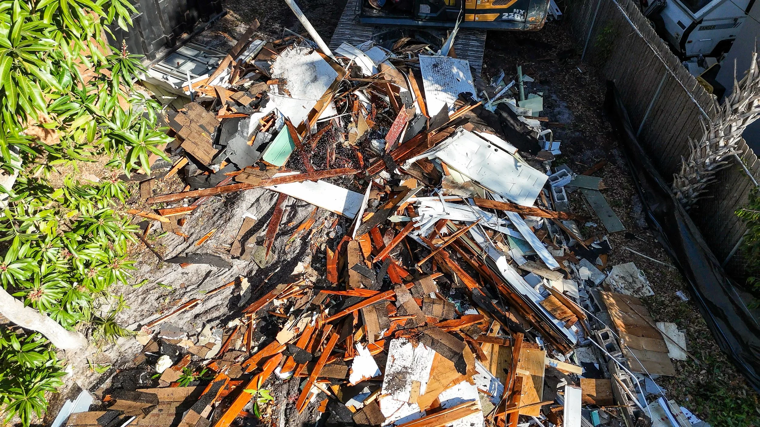 Image showing the debris and destruction after a fire, with charred wood, burnt furniture, and damaged household items in a backyard, surrounded by a wooden fence and some greenery.