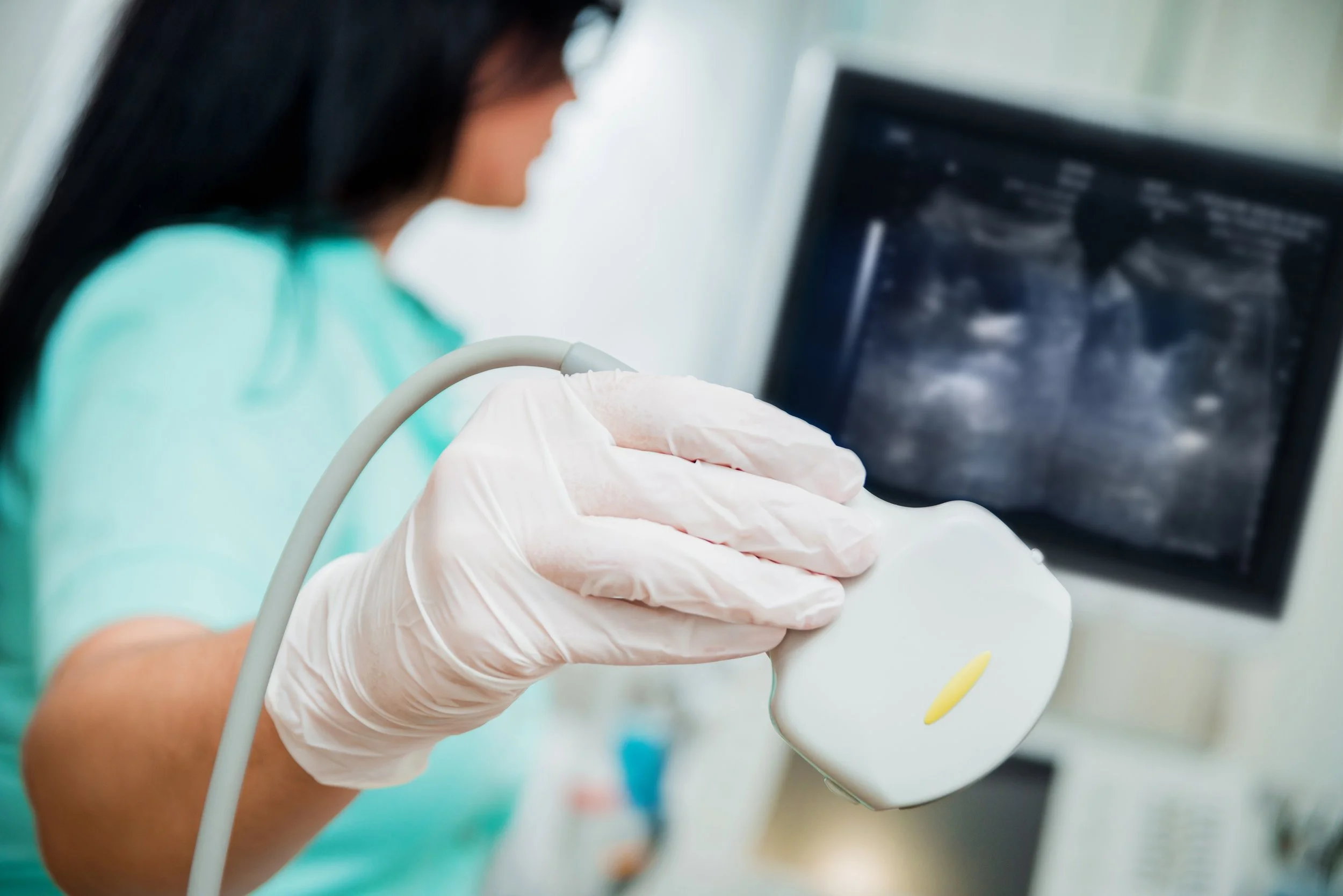 A healthcare professional wearing a green scrubs and white gloves holding an ultrasound transducer near a computer monitor showing an ultrasound image.