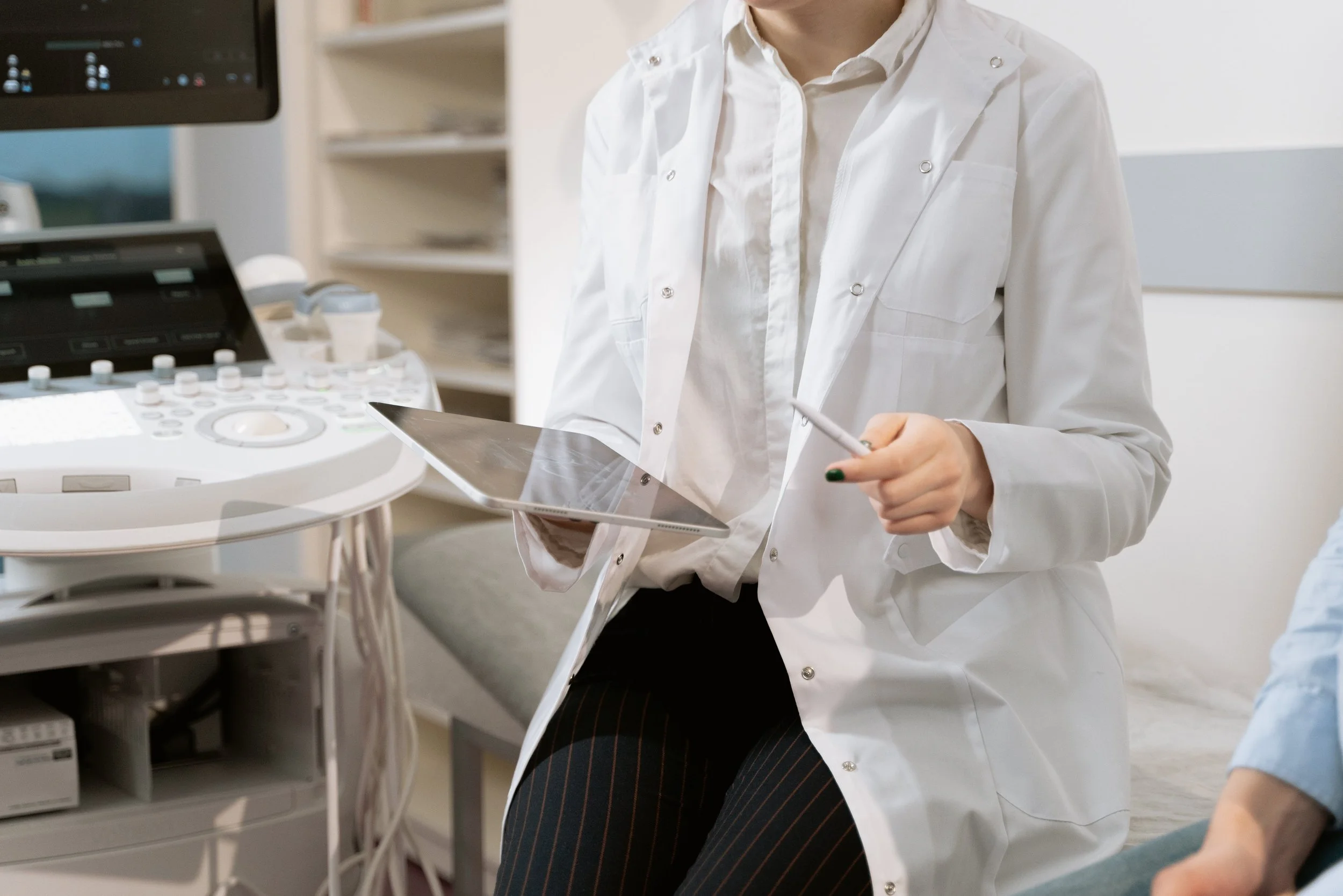 Medical professional in a white lab coat holding a tablet, standing next to medical equipment in a clinical setting.