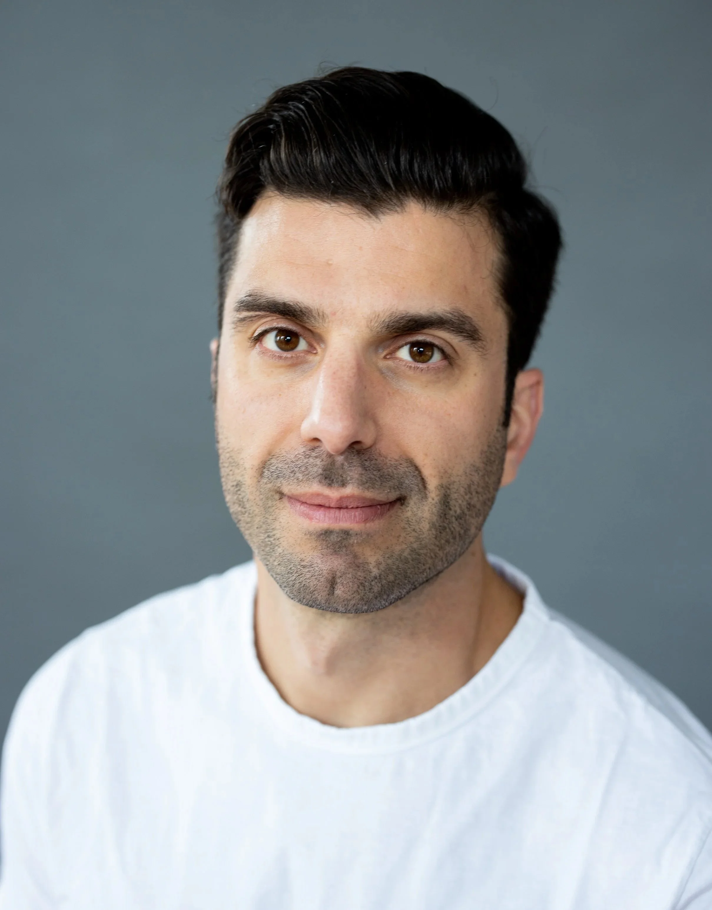 Close-up portrait of a young man with dark hair, brown eyes, and light skin, wearing a white shirt, against a gray background.