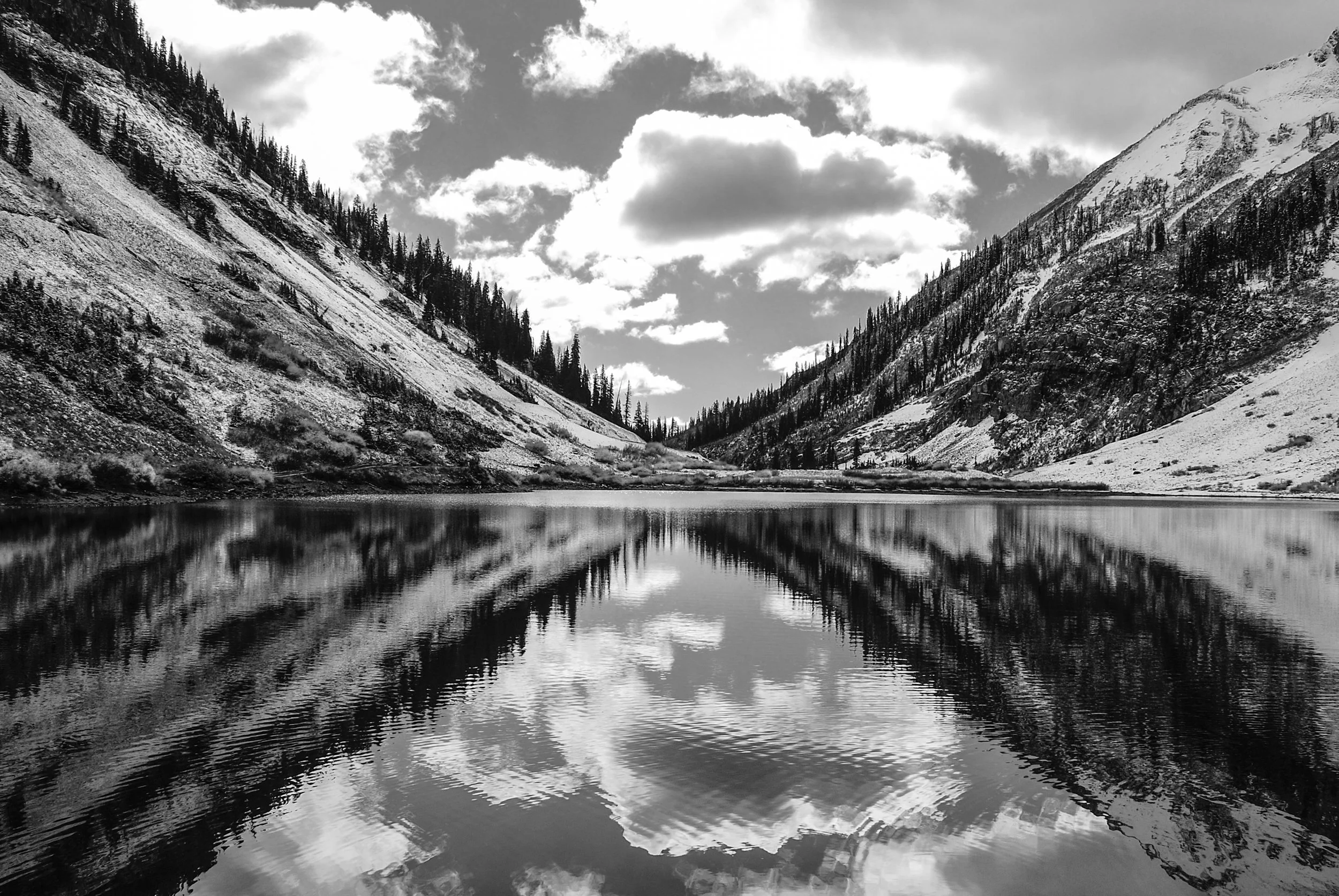 Black and white photograph of a mountain valley with snow-covered slopes, pine trees, and a partially cloudy sky. A calm lake reflects the surrounding landscape.