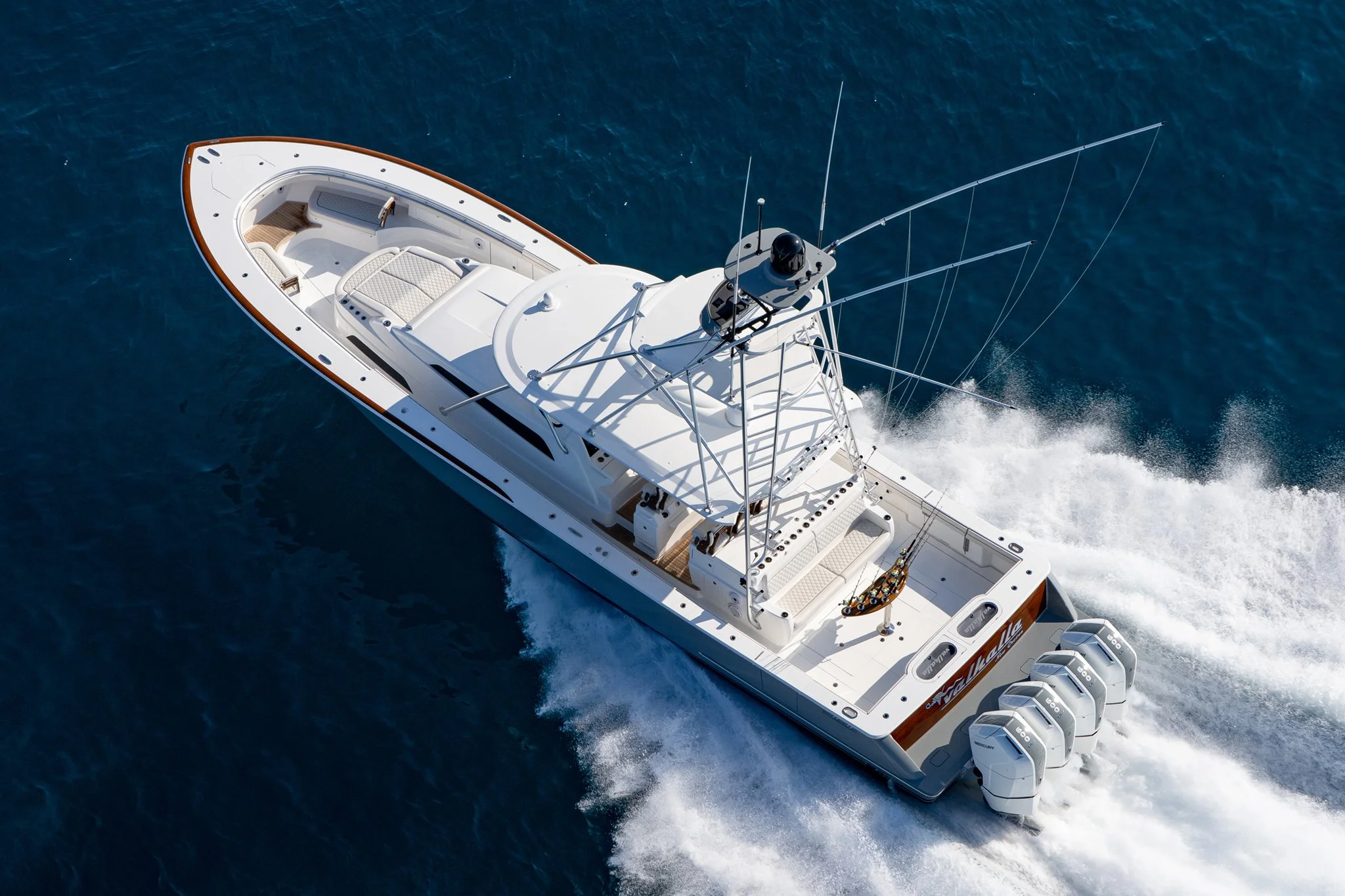 An aerial view of a white yacht speeding through blue ocean water, with four outboard engines at the stern and a shaded seating area on the deck.