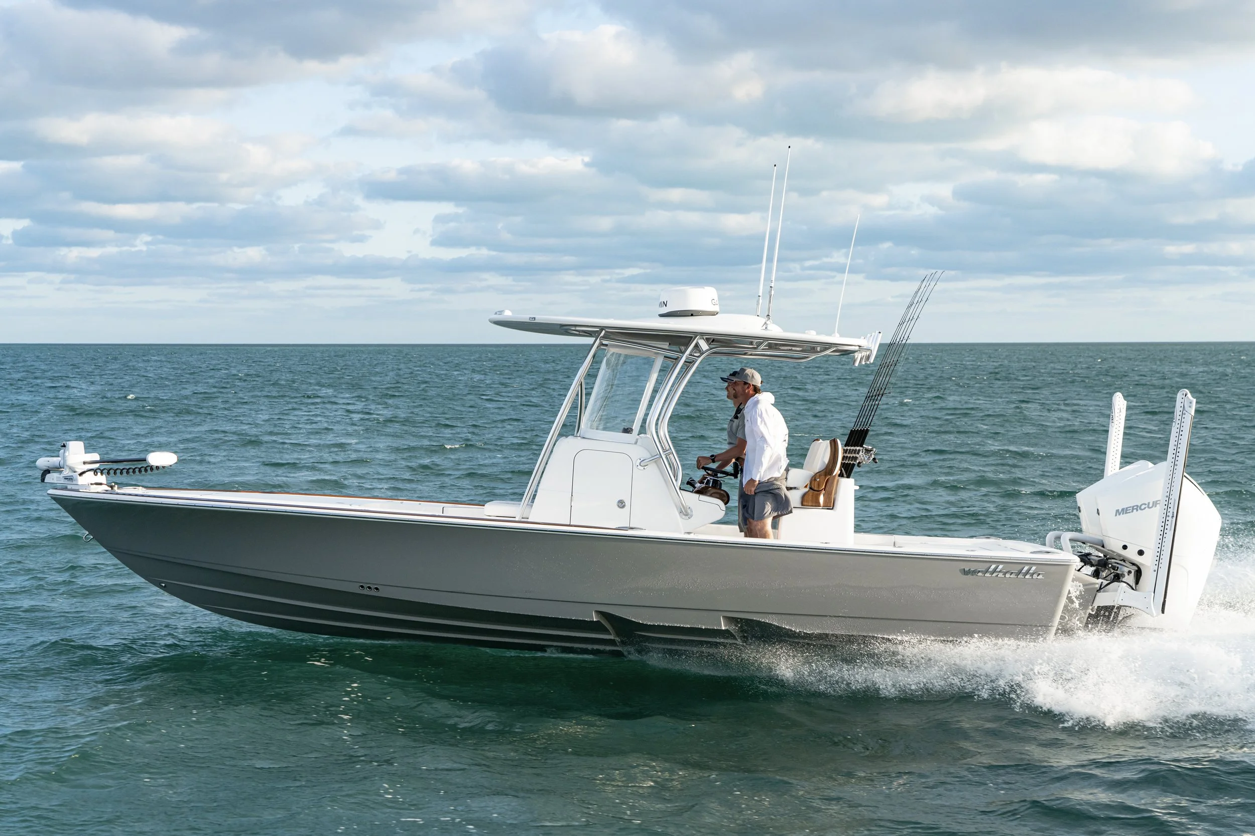 A man steering a white fishing boat with fishing rods on the side, moving over the water with a cloudy sky in the background.