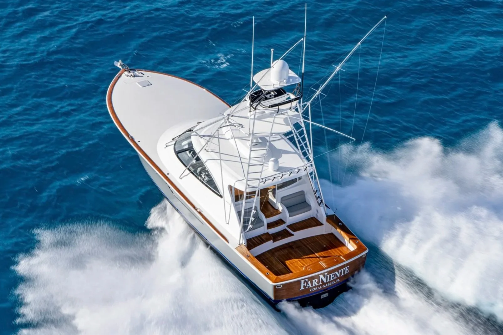 A white yacht named 'Far Niente' sailing on blue ocean water, creating wake behind it, with a wooden deck and boat equipment visible.