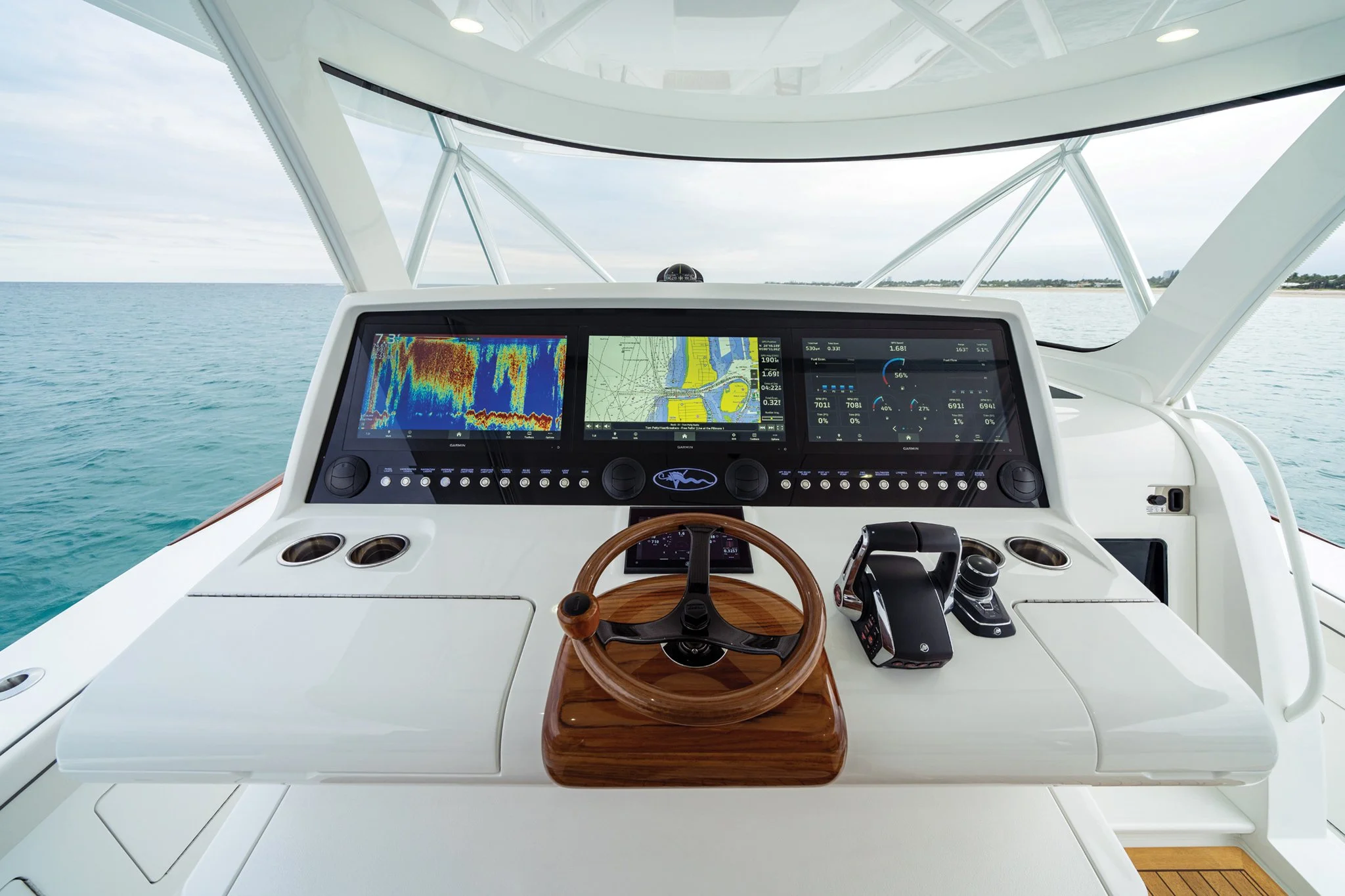 Overhead view of a yacht's helm station with multi-screen digital navigation and control systems, a wooden steering wheel, and a view of the ocean and shoreline through the windows.