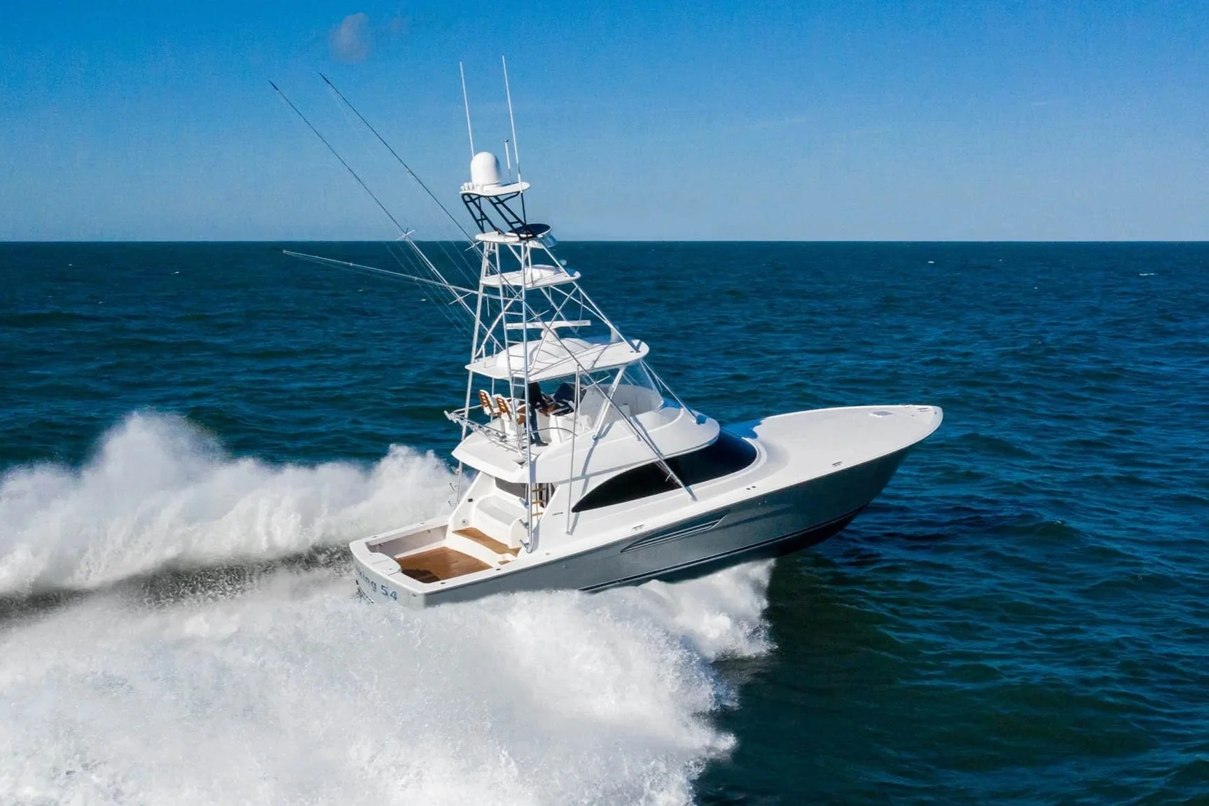 A white yacht with multiple decks and antennas is speeding through the ocean, creating waves and spray behind it under a clear blue sky.