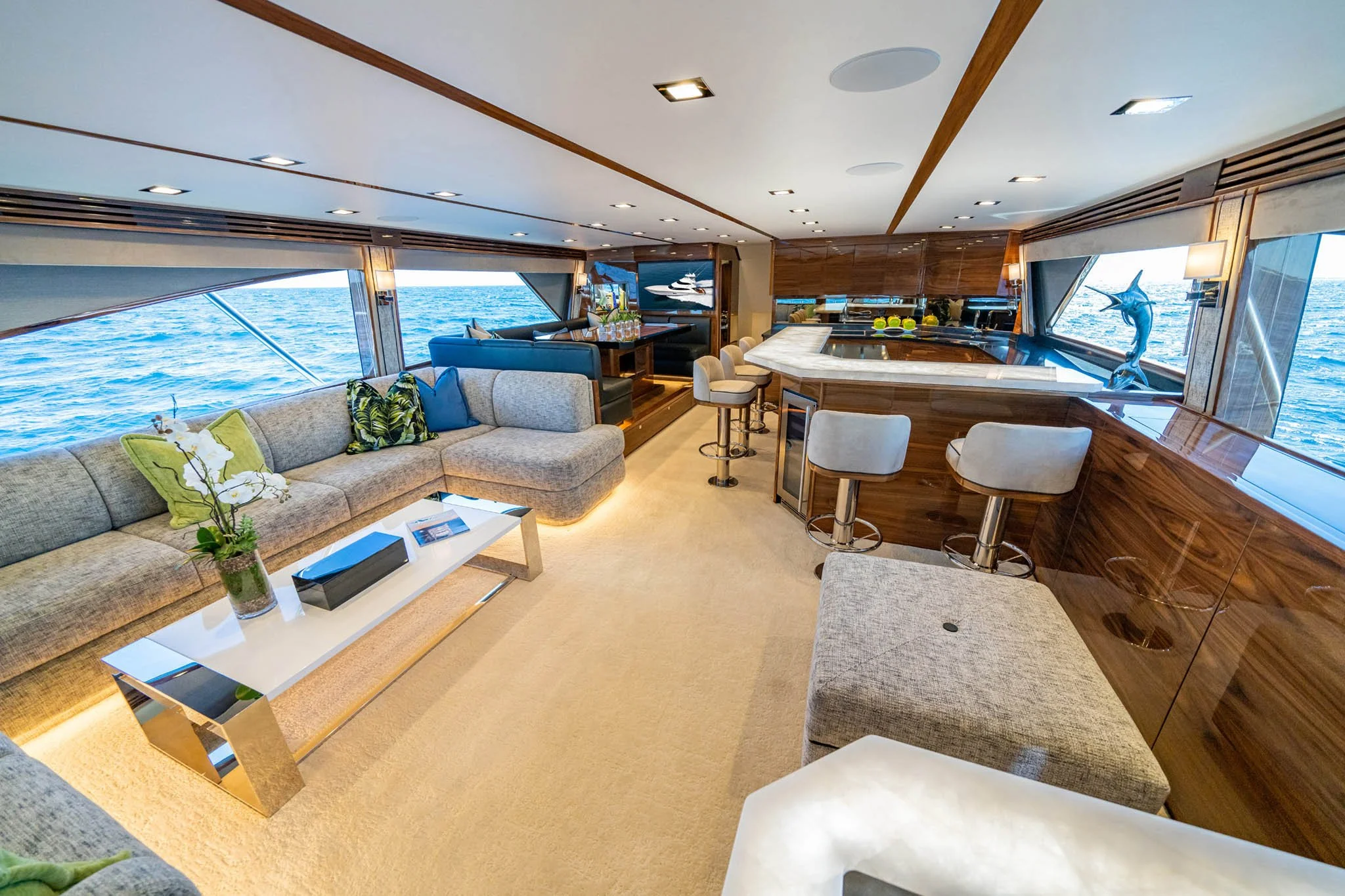 Interior of a yacht lounge with large windows showing the ocean, beige carpet, sectional sofa with green and blue pillows, a white coffee table with a plant, bar area with white bar stools, and decorative sculptures of marlin and fish.