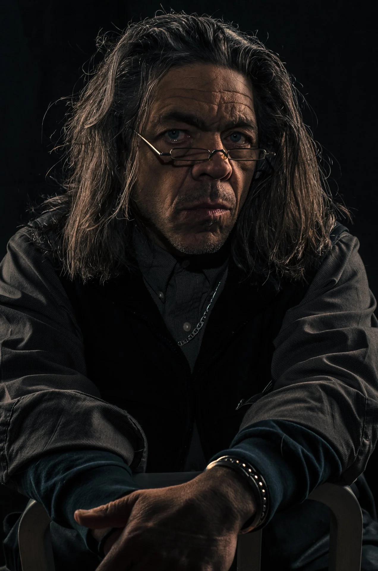 A middle-aged man with long, wavy gray hair and glasses perched on his nose, sitting with arms crossed in a dark setting.